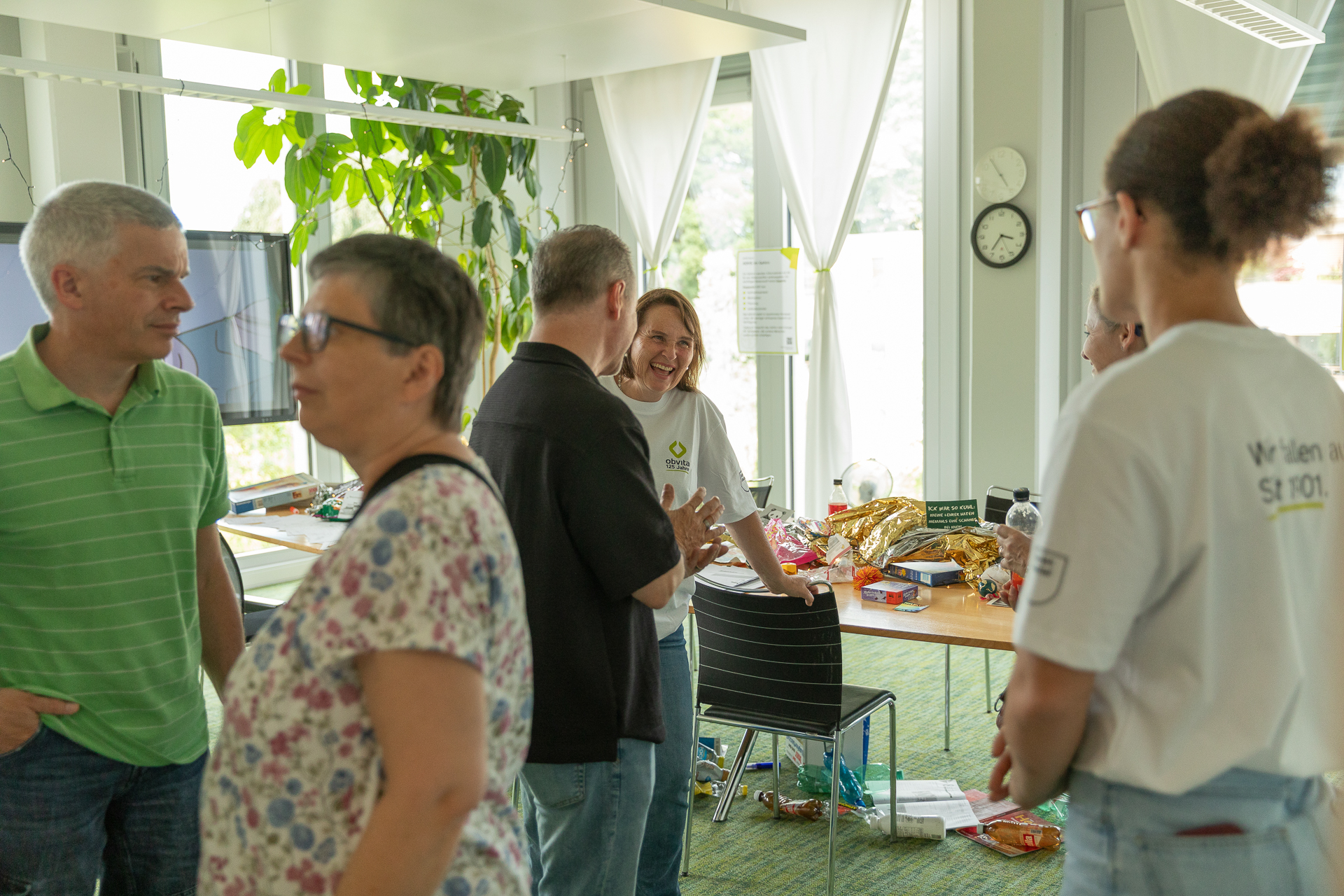 In einem hellen Raum mit großen Fenstern steht eine Gruppe von Menschen und unterhält sich. Im Hintergrund steht ein Tisch mit Snacks und Getränken. Einige tragen T-Shirts und Namensschilder, und die Atmosphäre wirkt ungezwungen und freundlich.