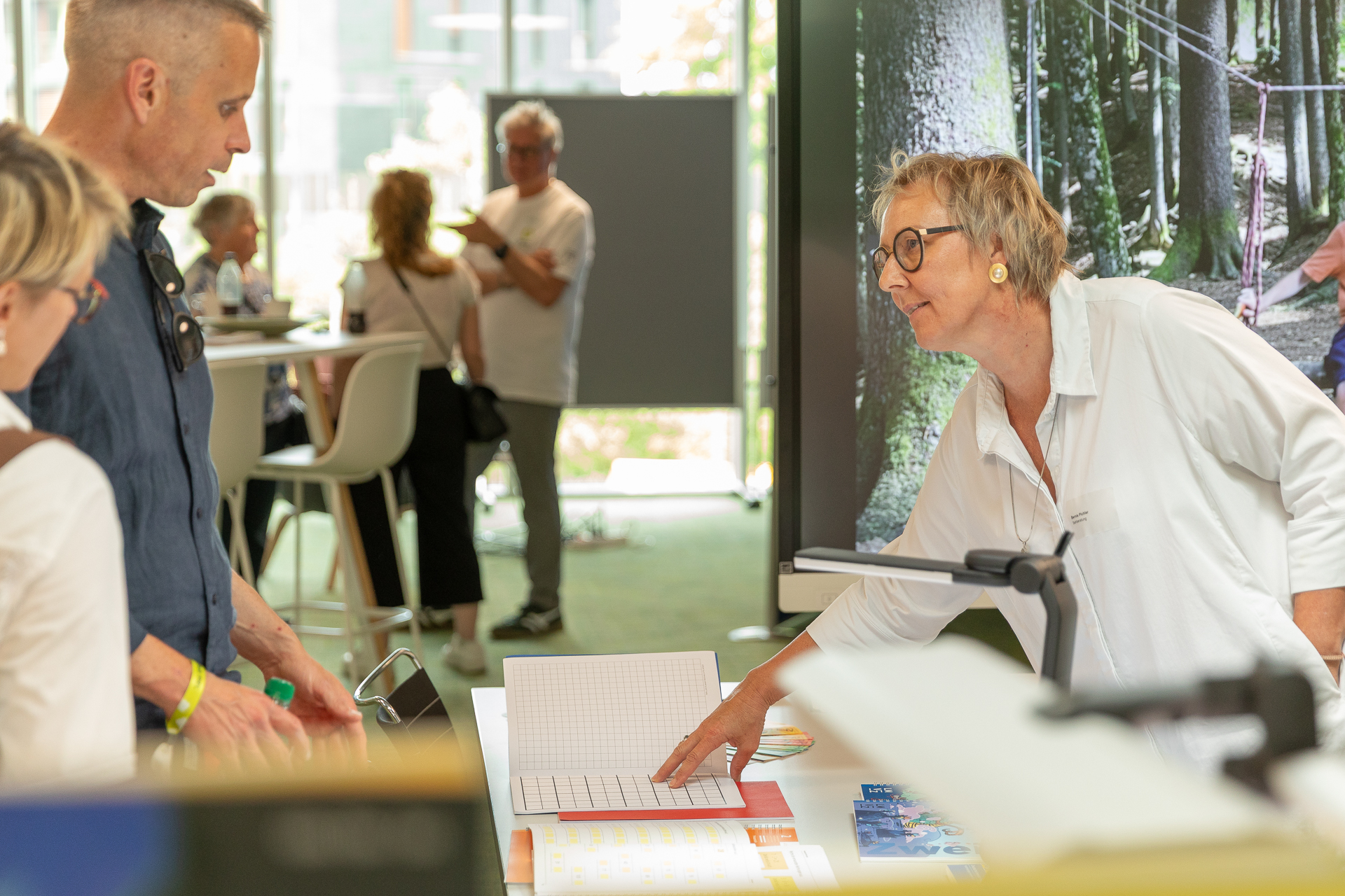 Eine Frau in einem weißen Hemd beugt sich über einen Tisch und zeigt zwei Personen bei einer Veranstaltung in einem Saal ein Notizbuch. Im Hintergrund sind weitere Personen zu sehen, und in der Nähe hängen große Plakate.