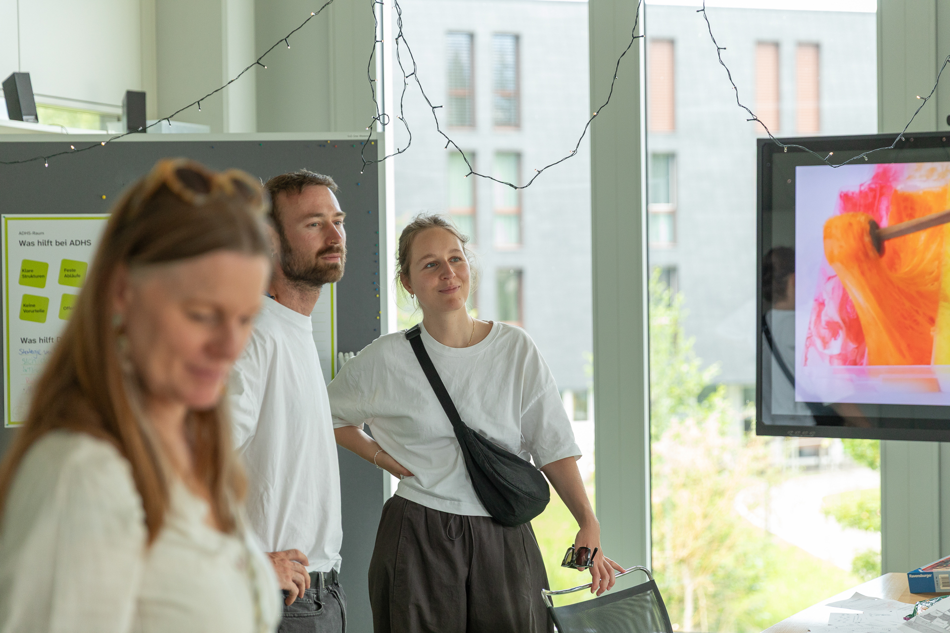 Drei Personen stehen in einem hellen Raum mit großen Fenstern. Zwei von ihnen blicken auf einen Bildschirm, auf dem farbenfrohe abstrakte Kunst zu sehen ist, während eine Frau im Vordergrund leicht unscharf abgebildet ist. Über ihnen hängen Lichterketten, und an den Wänden hängen Plakate.