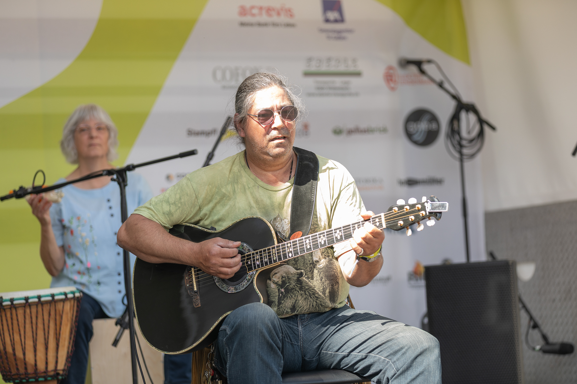 Ein Mann mit Sonnenbrille spielt auf einer Außenbühne Akustikgitarre, während im Hintergrund eine Frau neben einem Schlagzeug steht. Im Hintergrund sind verschiedene Sponsorenlogos zu sehen. Beide scheinen ganz in die Musik vertieft zu sein.