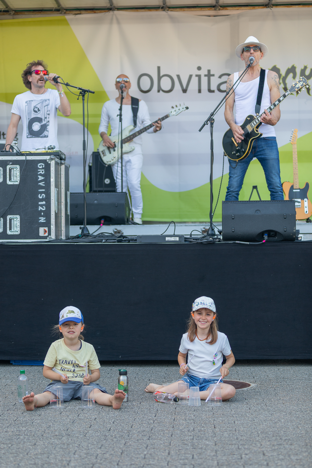 Zwei Kinder sitzen barfuß auf dem Boden und lächeln vor einer Bühne, während sie Getränke in den Händen halten. Hinter ihnen spielt eine Band mit vier Musikern in Freizeitkleidung und Hüten mit Instrumenten und Mikrofonen.