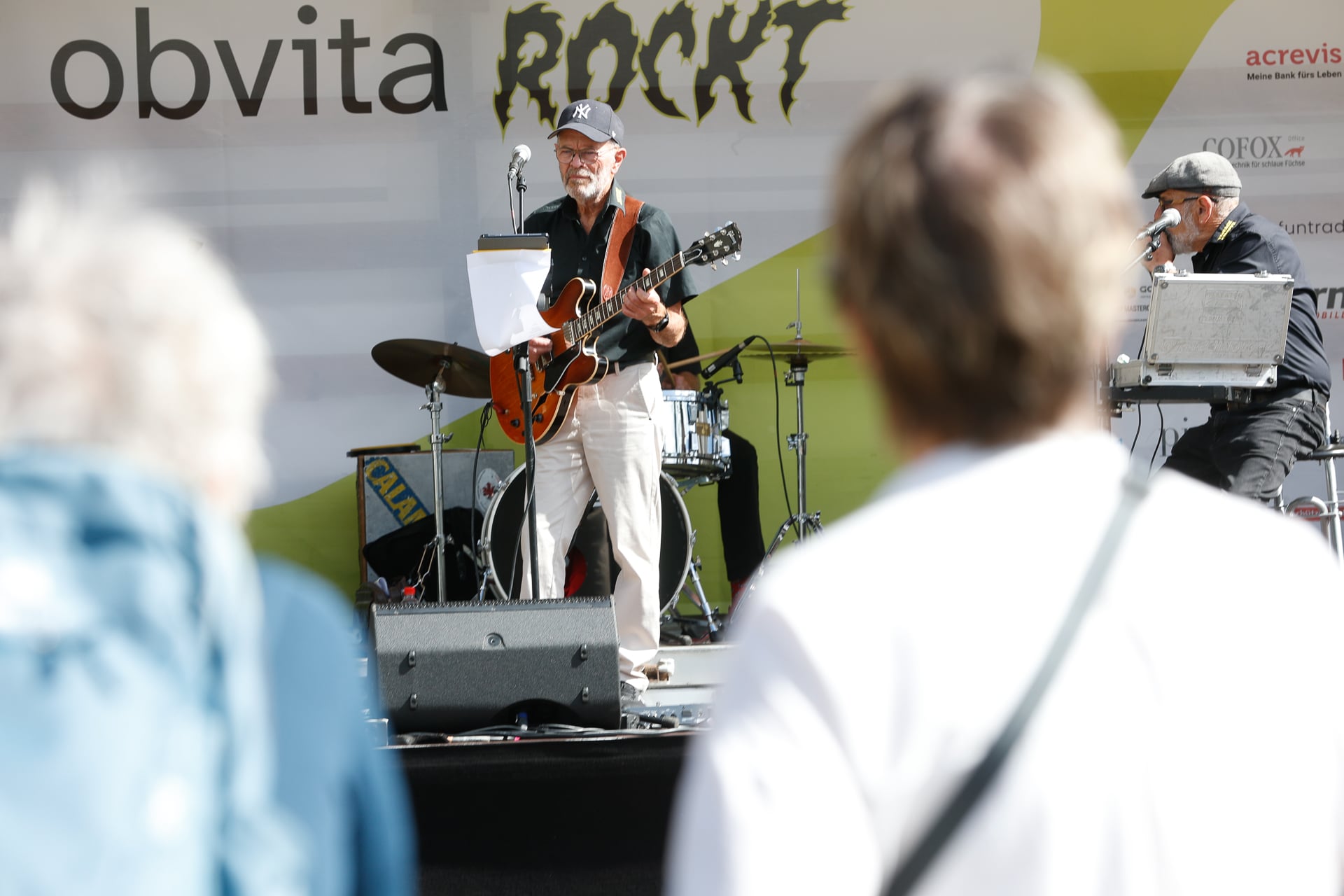 Ein Mann mit Mütze und Brille spielt auf einer Außenbühne E-Gitarre und singt, hinter ihm steht ein Schlagzeug, während die Menschen im Vordergrund der Darbietung zuschauen. Auf dem Banner auf der Bühne ist das Wort „ROCKT“ zu sehen.