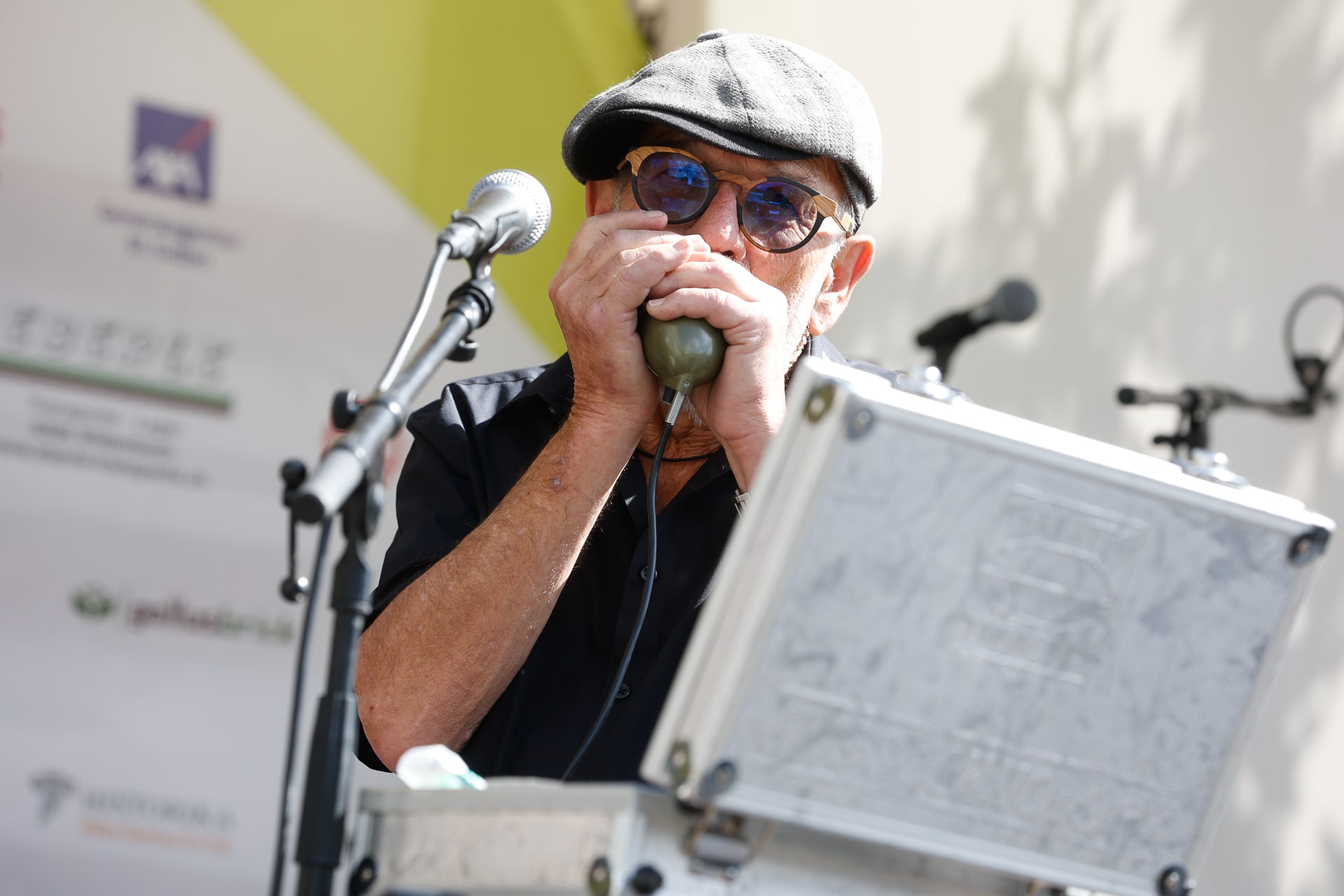 Ein Mann mit Sonnenbrille, Schirmmütze und dunklem Hemd spielt Mundharmonika in ein Mikrofon. Er steht hinter einem Metallkoffer, hinter ihm sind verschwommene Mikrofone und ein mit Logos übersäter Hintergrund zu sehen.
