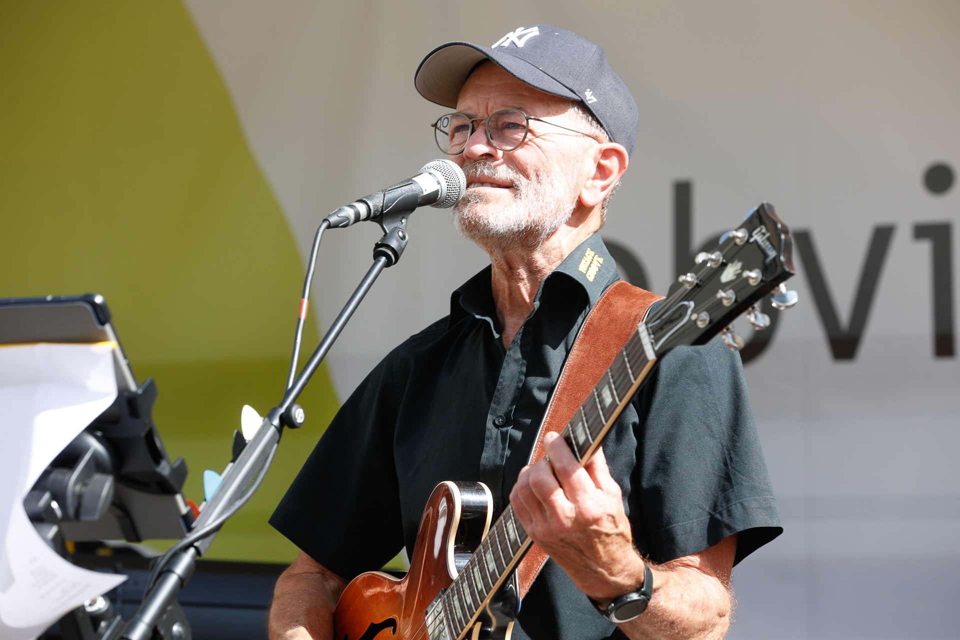 Ein älterer Mann mit Brille, Mütze und schwarzem Hemd singt bei einer Veranstaltung im Freien in ein Mikrofon, während er E-Gitarre spielt. Vor ihm ist ein Notenständer mit Notenblättern zu sehen.
