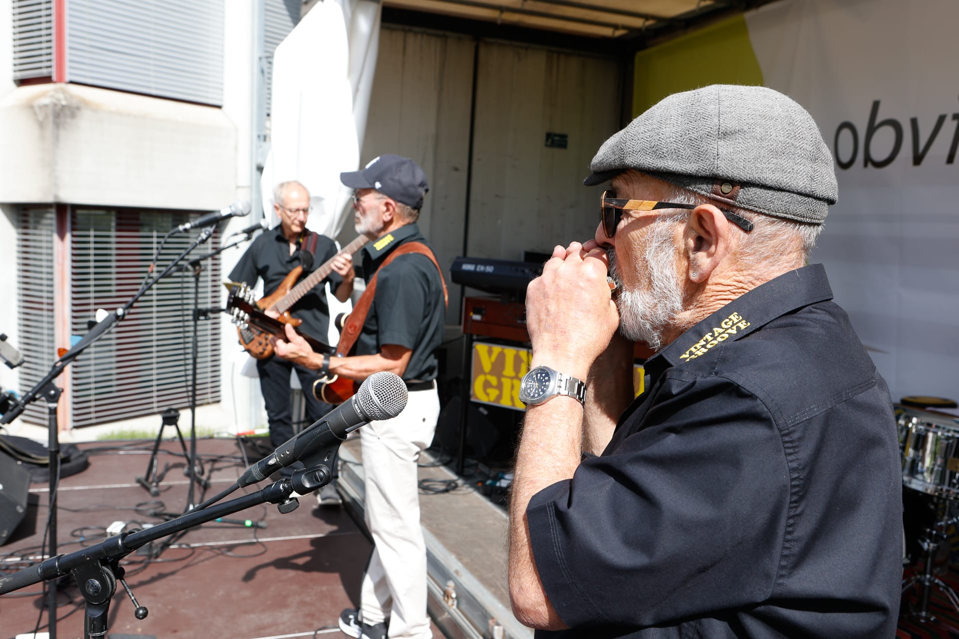 Drei ältere Männer spielen auf einer Bühne im Freien; einer spielt Gitarre, ein anderer Bass und der dritte, der eine Mütze trägt, spielt Mundharmonika. Um sie herum sind Mikrofone und Instrumente aufgestellt.