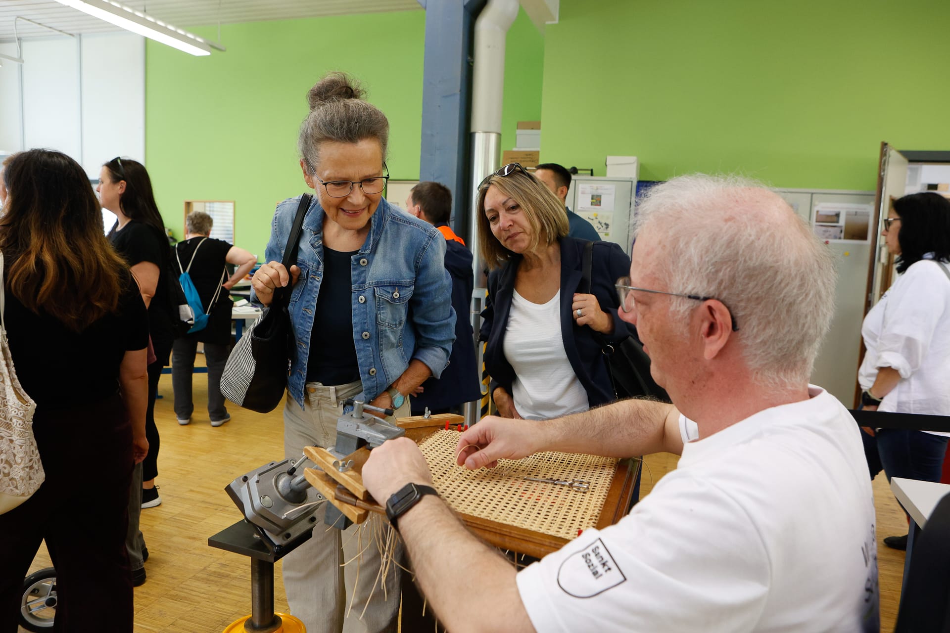 Ein Mann führt zwei interessierten Frauen in einem belebten Innenraum die traditionelle Stuhlflechttechnik vor, während andere im Hintergrund zuschauen und sich unterhalten.