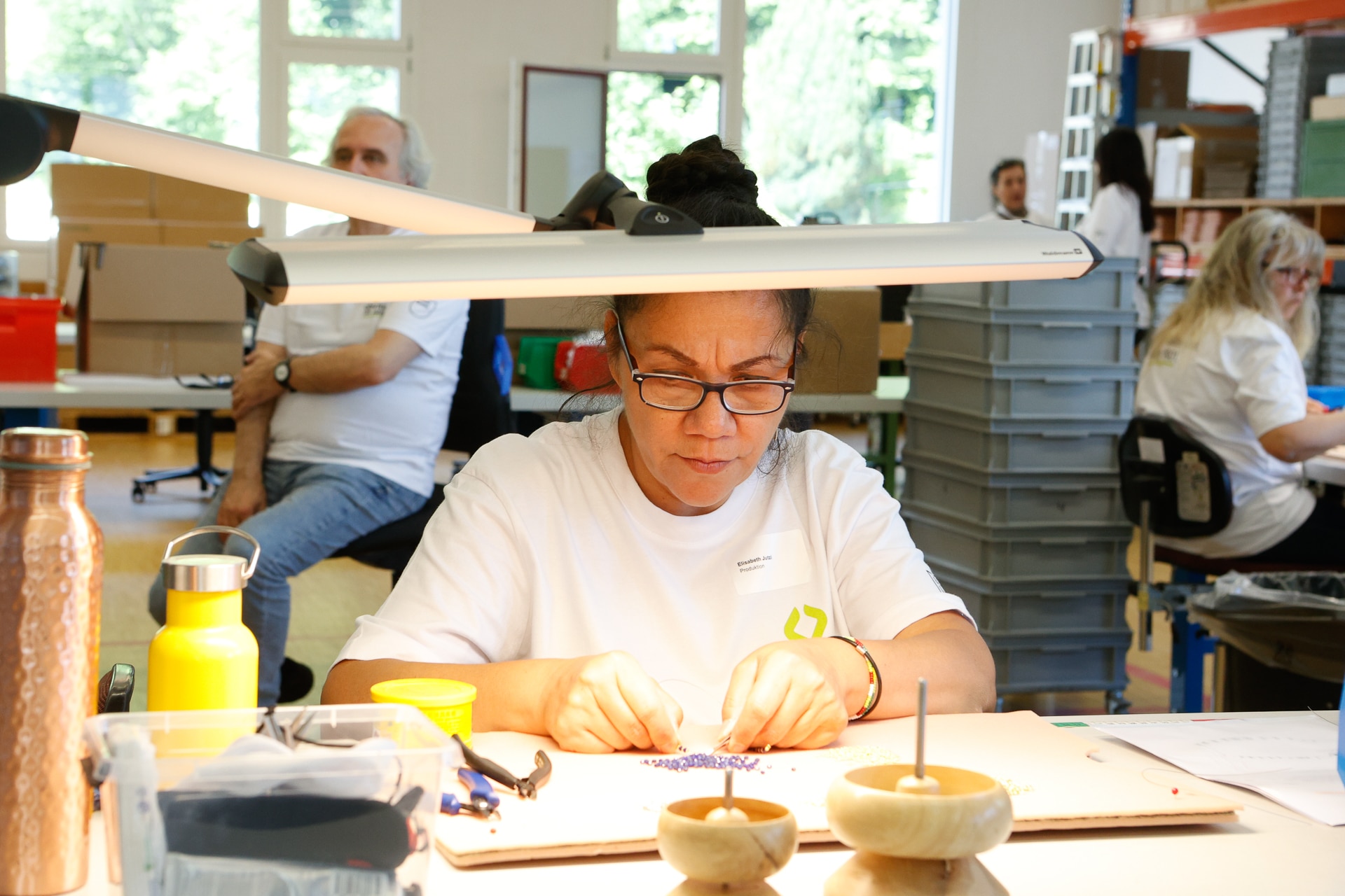Eine Frau mit Brille und in einem weißen Hemd arbeitet konzentriert an einem gut beleuchteten Tisch in einer Werkstatt an kleinen Werkstücken. Auf dem Tisch stehen Wasserflaschen und Werkzeuge, im Hintergrund sind weitere Personen bei der Arbeit zu sehen.