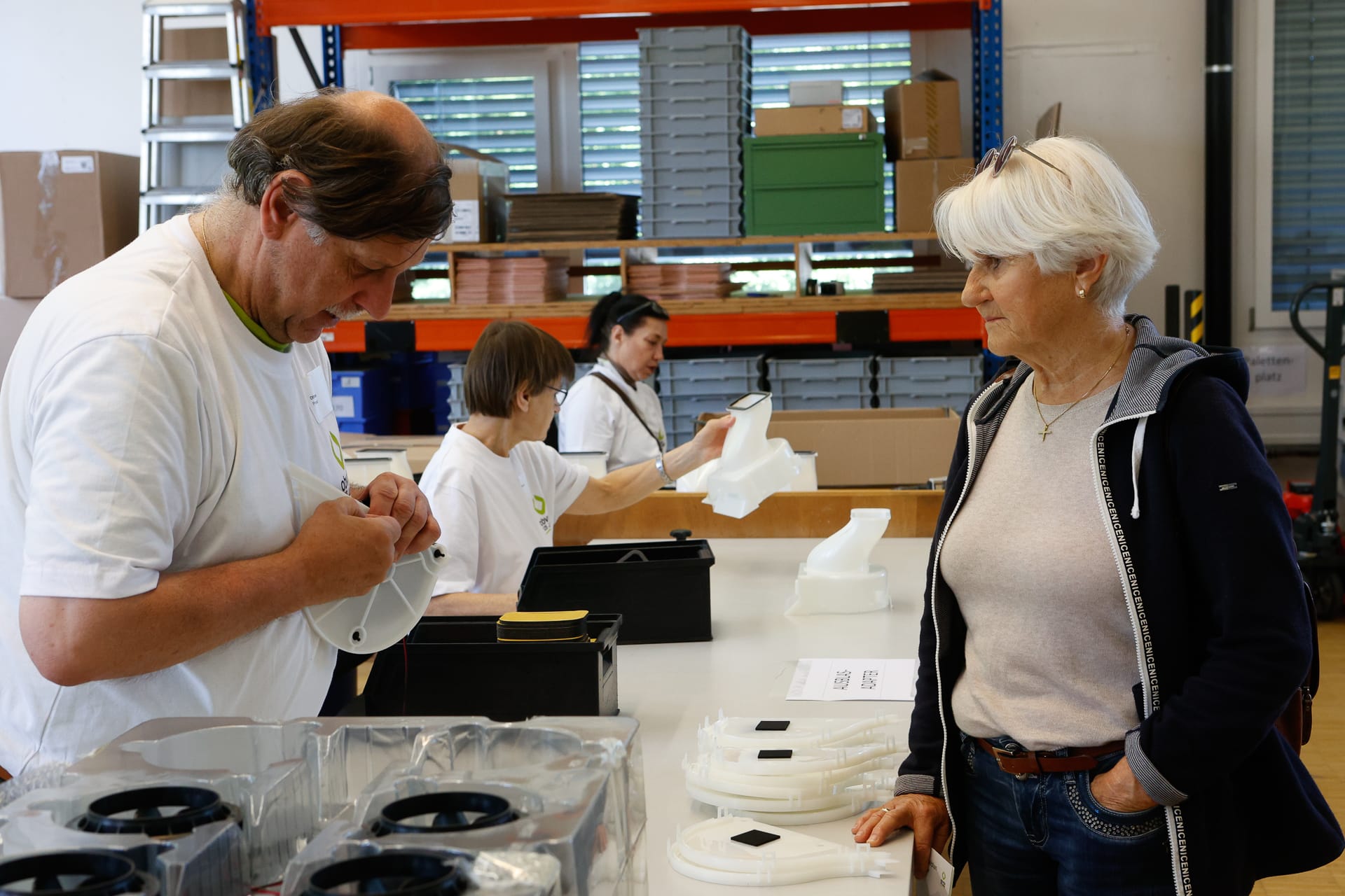 Zwei Frauen und ein Mann arbeiten gemeinsam in einer Montagehalle. Ein Mann beschriftet ein weißes Teil, während eine Frau dabei zusieht. Im Hintergrund sind Regale mit Kisten und Materialien zu sehen.