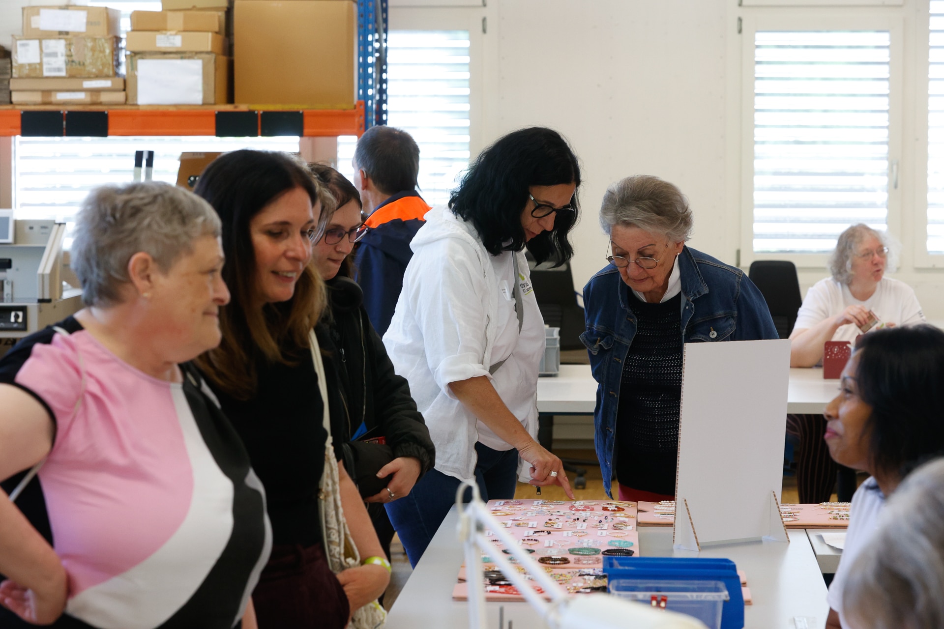 Eine Gruppe von Frauen steht in einem Raum um einen Tisch herum und betrachtet Gegenstände, die zum Verkauf oder zur Ausstellung ausgelegt sind. Einige unterhalten sich; eine Frau zeigt auf etwas auf dem Tisch. Im Hintergrund des Raums sind Regale, Kisten und Fenster zu sehen.