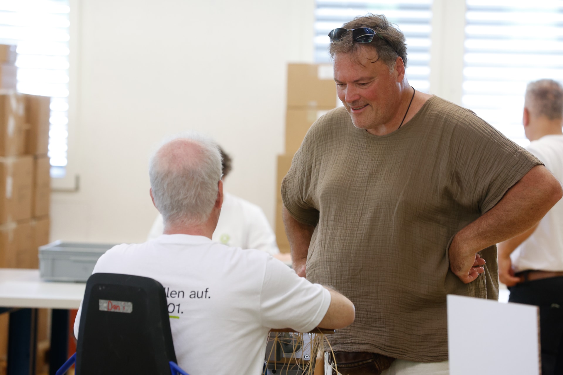 Zwei Männer unterhalten sich in einem hellen Raum, im Hintergrund stehen Pappkartons; einer sitzt mit dem Rücken zur Kamera, während der andere ihm gegenüber steht, die Hände in die Hüften gestemmt, und lächelt.