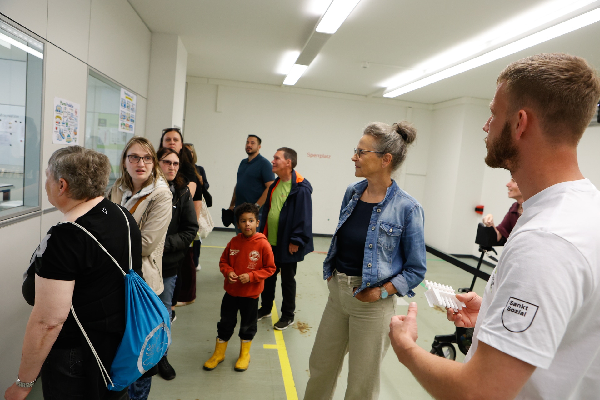 Eine Gruppe aus Erwachsenen und Kindern steht in einem weißen Raum; einige betrachten Plakate an der Wand, während ein Mann in einem weißen Hemd einen kleinen Gegenstand in der Hand hält und ihn den anderen zeigt. Die Atmosphäre wirkt lehrreich oder wie bei einer Führung.
