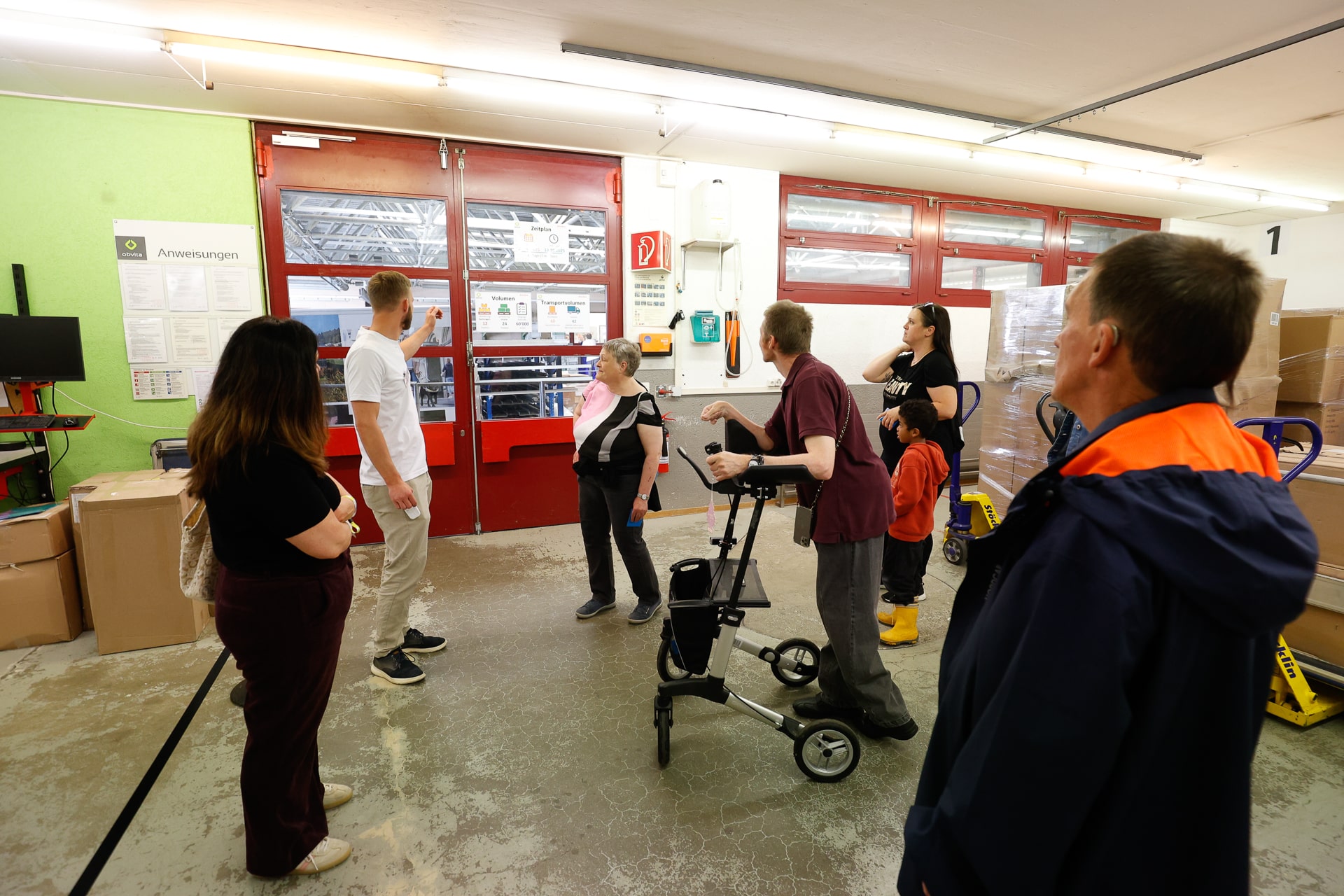 Eine Gruppe von Menschen, darunter eine Person mit einem Rollator, steht in einem hell beleuchteten Innenraum mit roten Türen, Kisten und Paletten und scheint einem Führer zuzuhören oder Anweisungen zu erhalten.