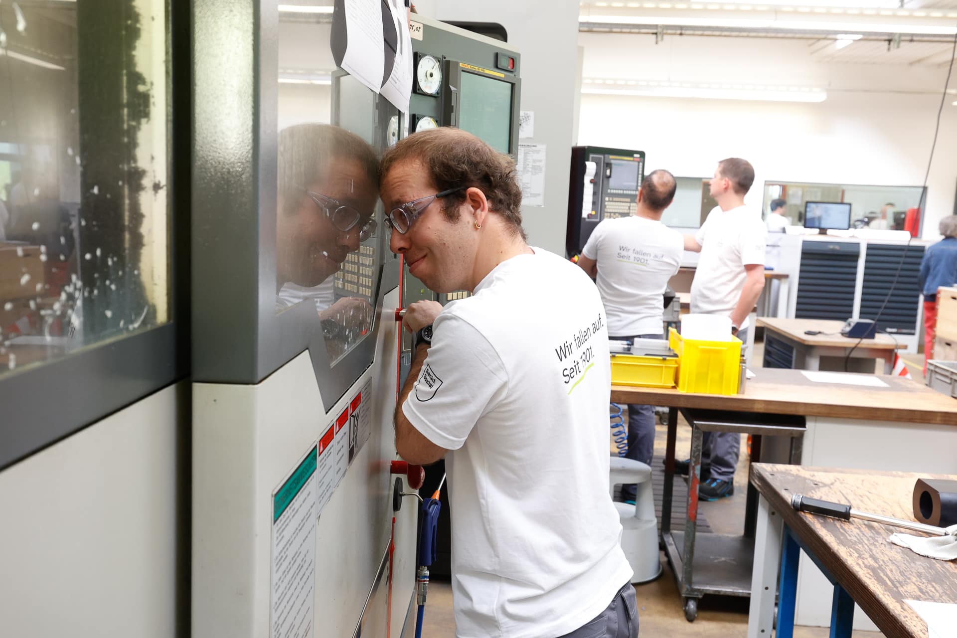 Ein Mann mit Schutzbrille bedient eine Maschine in einer hellen, modernen Werkstatt. Sein Spiegelbild ist in der Glasscheibe der Maschine zu sehen. Im Hintergrund arbeiten weitere Mitarbeiter in weißen Hemden an verschiedenen Maschinen.