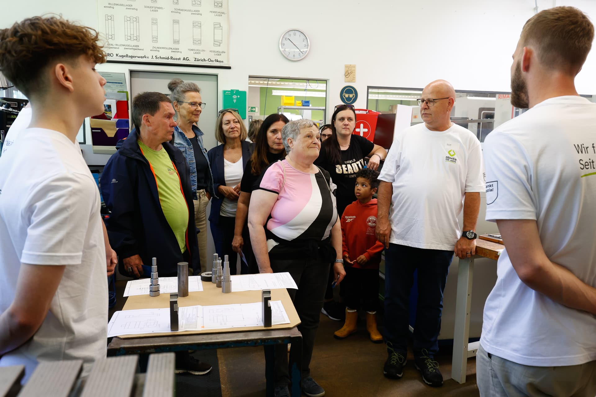 Eine Gruppe aus Erwachsenen und Kindern steht in einer Werkstatt und hört zwei Ausbildern in weißen Hemden zu. Man sieht Tische mit Papieren, Werkzeugen und Metallteilen. Die Umgebung wirkt lehrreich, an der Wand hängen Diagramme und eine Uhr.