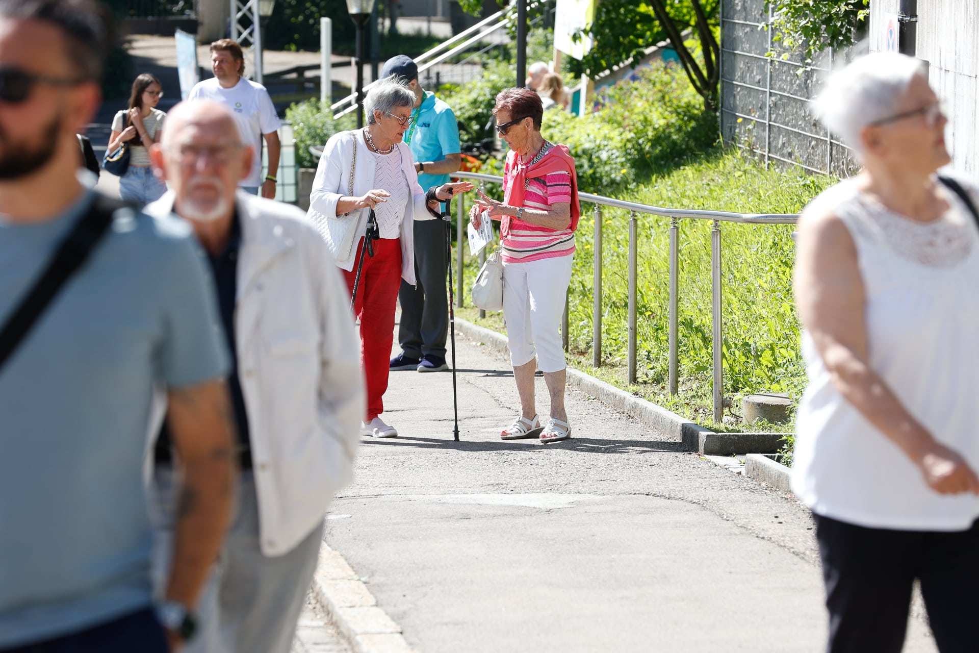 Eine Gruppe von Menschen, darunter ältere Frauen mit Gehstöcken, geht auf einem sonnigen, von Grün gesäumten Bürgersteig spazieren und unterhält sich, während im Vordergrund andere vorbeigehen.