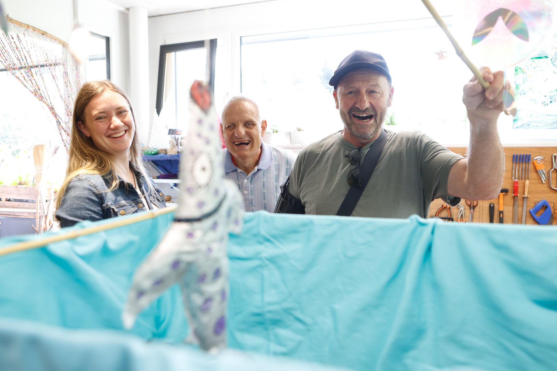 Drei Erwachsene lächeln und lachen, während sie einem verspielten Puppenspiel zusehen; dabei hält ein Mann hinter einem blauen Vorhang einen Stab über einer bunten Hundepuppe. Die Kulisse wirkt hell und fröhlich – es handelt sich um einen Innenraum.