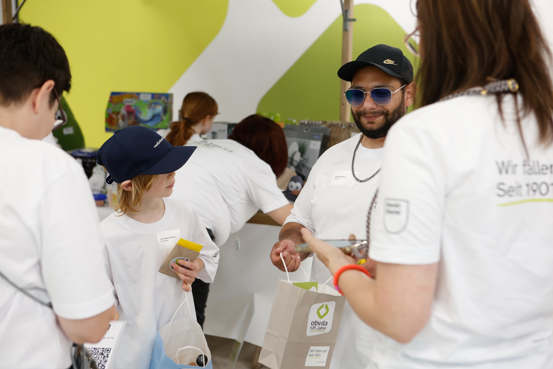 Eine Gruppe von Menschen in weißen Hemden und Mützen unterhält sich bei einer Veranstaltung in einem Saal, während sie Geschenktüten in den Händen halten. Eine Person lächelt und trägt eine Sonnenbrille sowie eine blaue Mütze; von den anderen sind nur die Rücken zu sehen. Im Hintergrund ist eine grün-weiße Wand zu sehen.