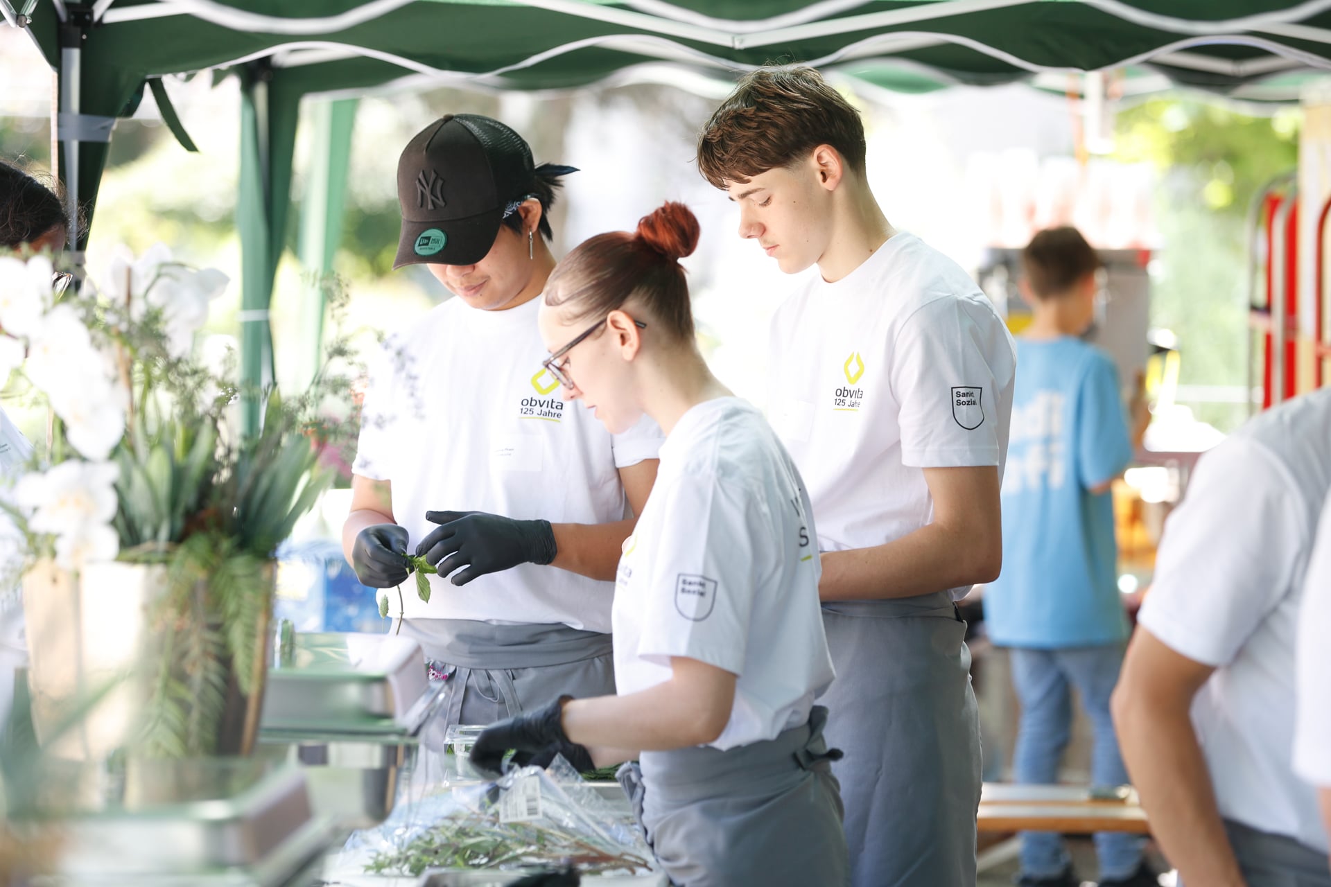 Drei junge Leute in weißen Hemden und grauen Schürzen bereiten gemeinsam im Freien unter einem Vordach Essen zu. Zwei von ihnen sind damit beschäftigt, Kräuter zu verarbeiten, während der Dritte zusieht. Im Hintergrund sind Blumen und verschwommene Menschen zu sehen.