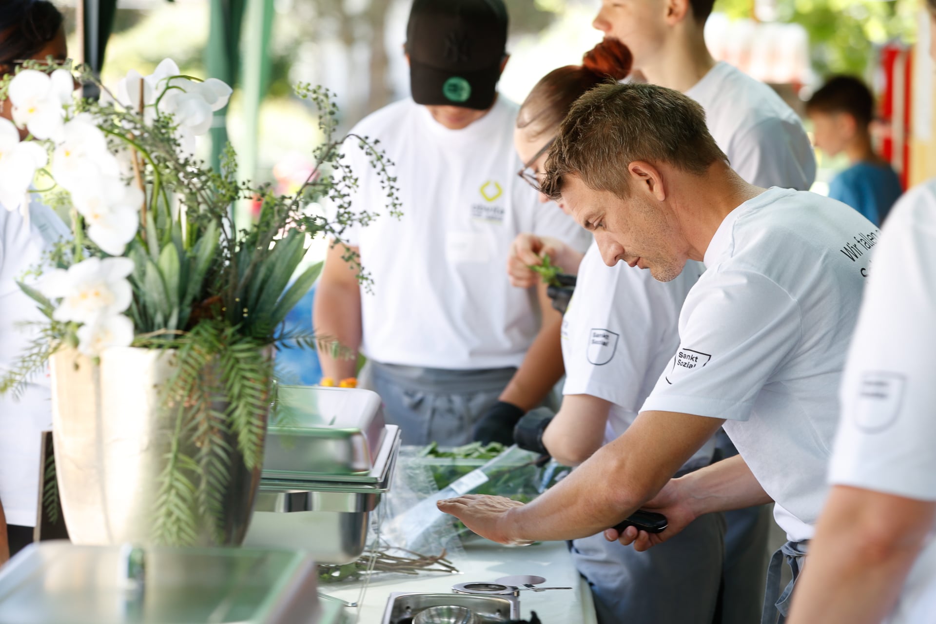 Eine Gruppe von Köchen in weißen Uniformen bereitet bei einer Veranstaltung im Freien Speisen zu und arbeitet gemeinsam an einem Tisch, der mit einer Vase voller weißer Blumen und Grünpflanzen geschmückt ist. Ein Koch hackt Kräuter, während andere ihm dabei helfen.