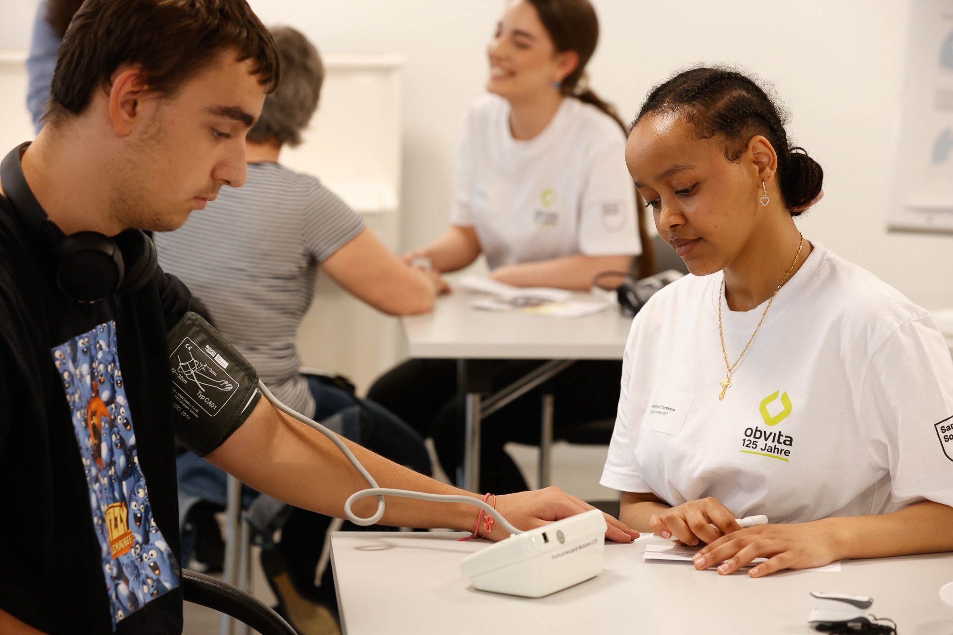 Eine Frau in einem weißen Hemd misst an einem Tisch mit einem digitalen Blutdruckmessgerät den Blutdruck eines jungen Mannes. Im Hintergrund sitzen weitere Personen und unterhalten sich. Der Schauplatz scheint eine Gesundheitsklinik oder eine Gemeinschaftsveranstaltung zu sein.