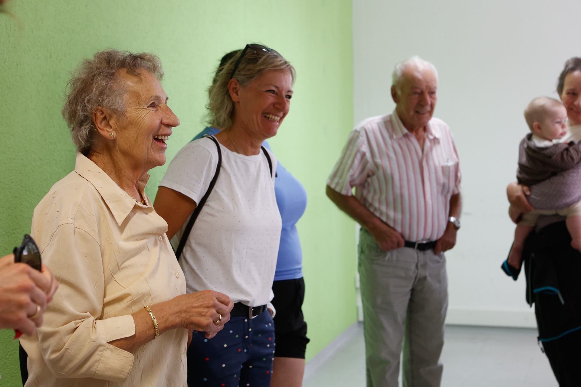 Eine Gruppe lächelnder Menschen, darunter zwei ältere Frauen, ein älterer Mann, eine jüngere Frau und ein Baby, steht gemeinsam in einem Raum vor einer grün-weißen Wand.