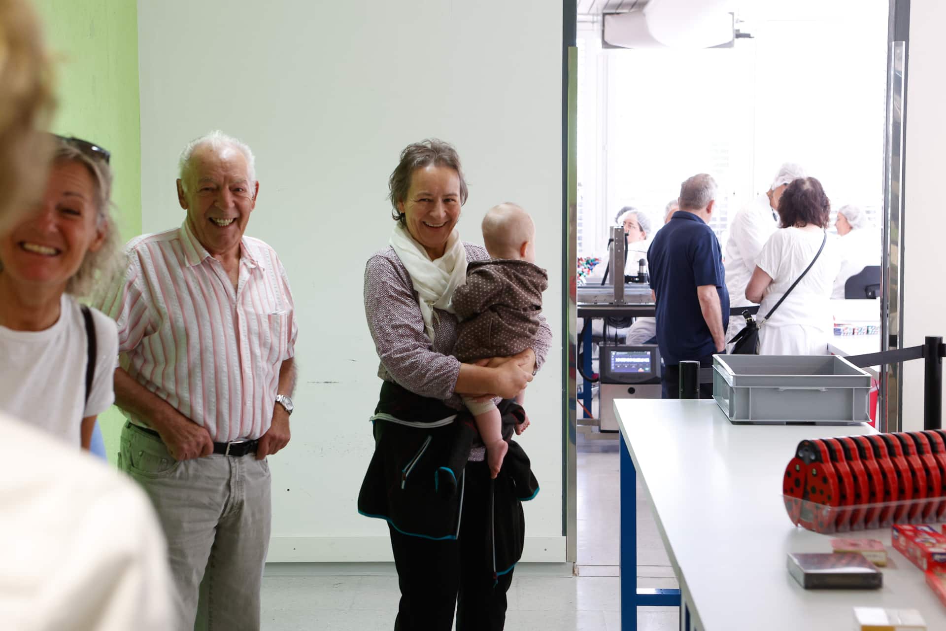 Ein lächelnder älterer Mann und eine ältere Frau, die ein Baby im Arm hält, stehen in einem hellen Raum, während im Hintergrund durch eine Tür weitere Personen zu sehen sind. Im Vordergrund stehen ein Tisch und verschiedene Geräte.