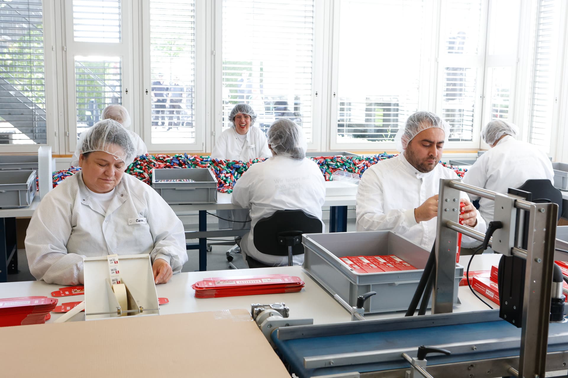Mitarbeiter in weißen Laborkitteln und Haarnetzen montieren und verpacken Produkte an einem hellen, sauberen Arbeitsplatz in einer Fabrik, im Hintergrund sind große Fenster und bunte Materialien zu sehen.