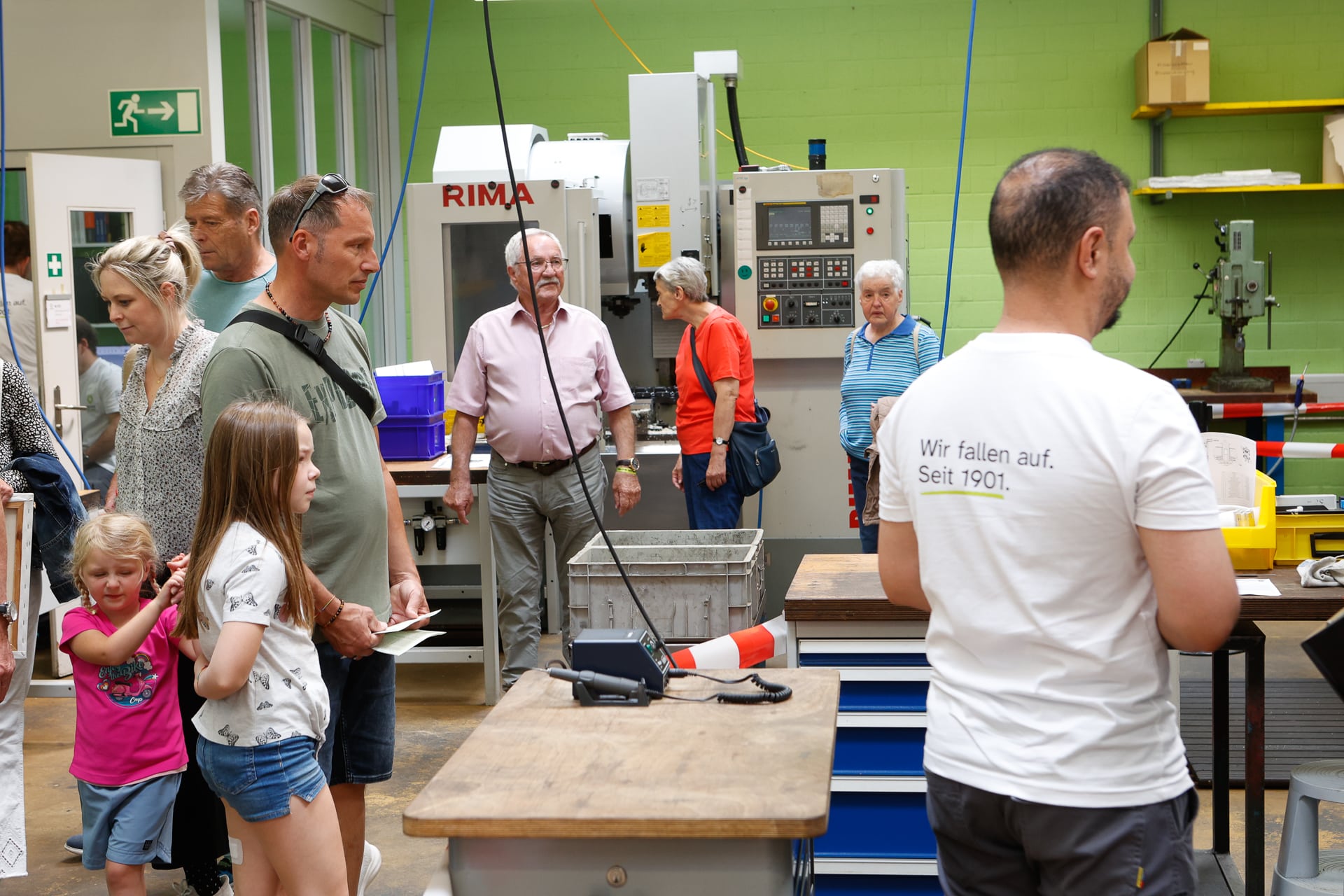 Eine Gruppe von Menschen, darunter Kinder und Erwachsene, steht in einer Werkstatt mit Maschinen und Werkbänken. Ein Mann, der von hinten zu sehen ist, trägt ein weißes Hemd mit deutscher Aufschrift. Die Atmosphäre ist ungezwungen und aufmerksam.