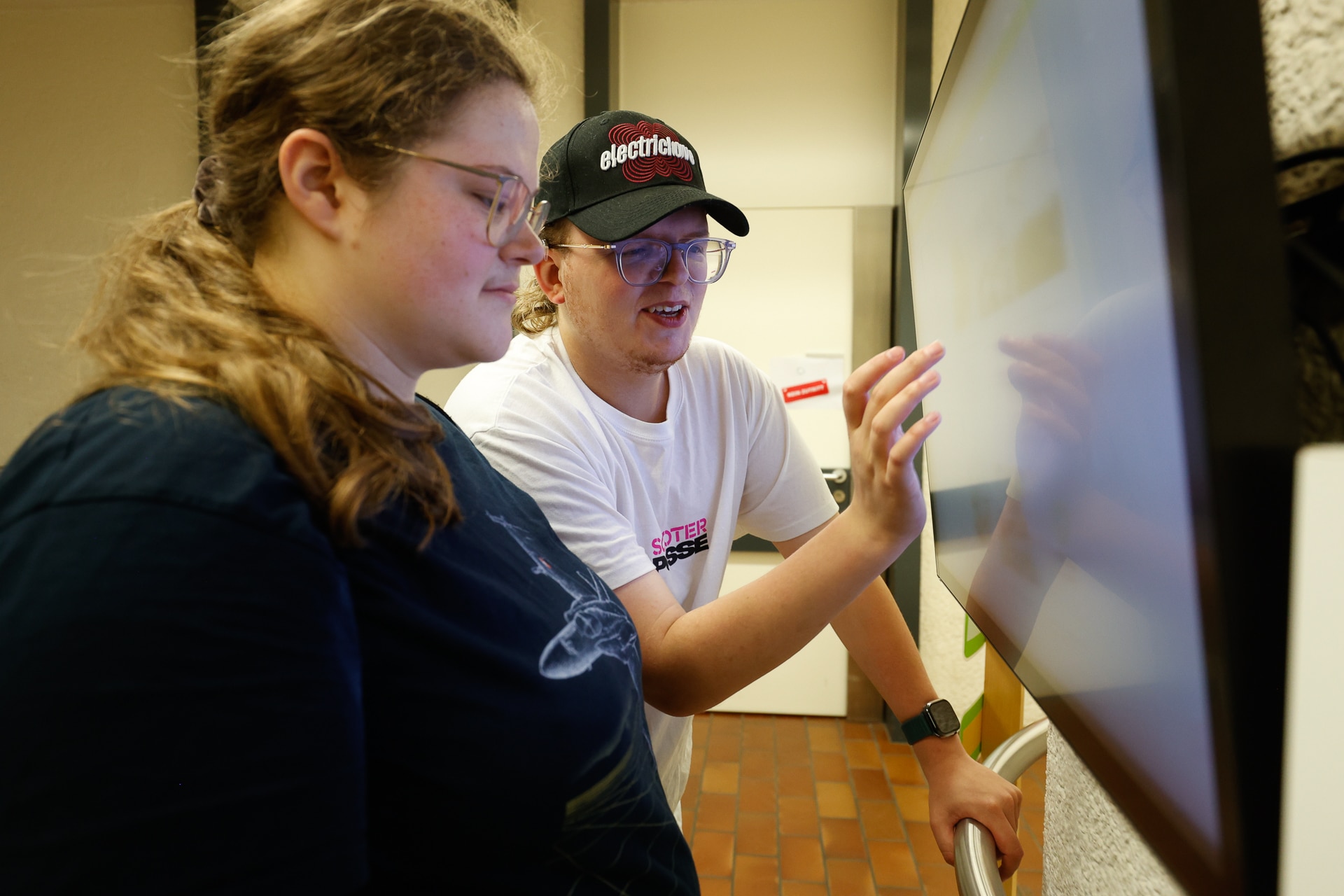 Zwei Personen stehen nebeneinander und bedienen einen großen Touchscreen. Eine Person zeigt auf den Bildschirm, während sie etwas erklärt, und die andere schaut aufmerksam zu. Beide tragen eine Brille und Freizeitkleidung.
