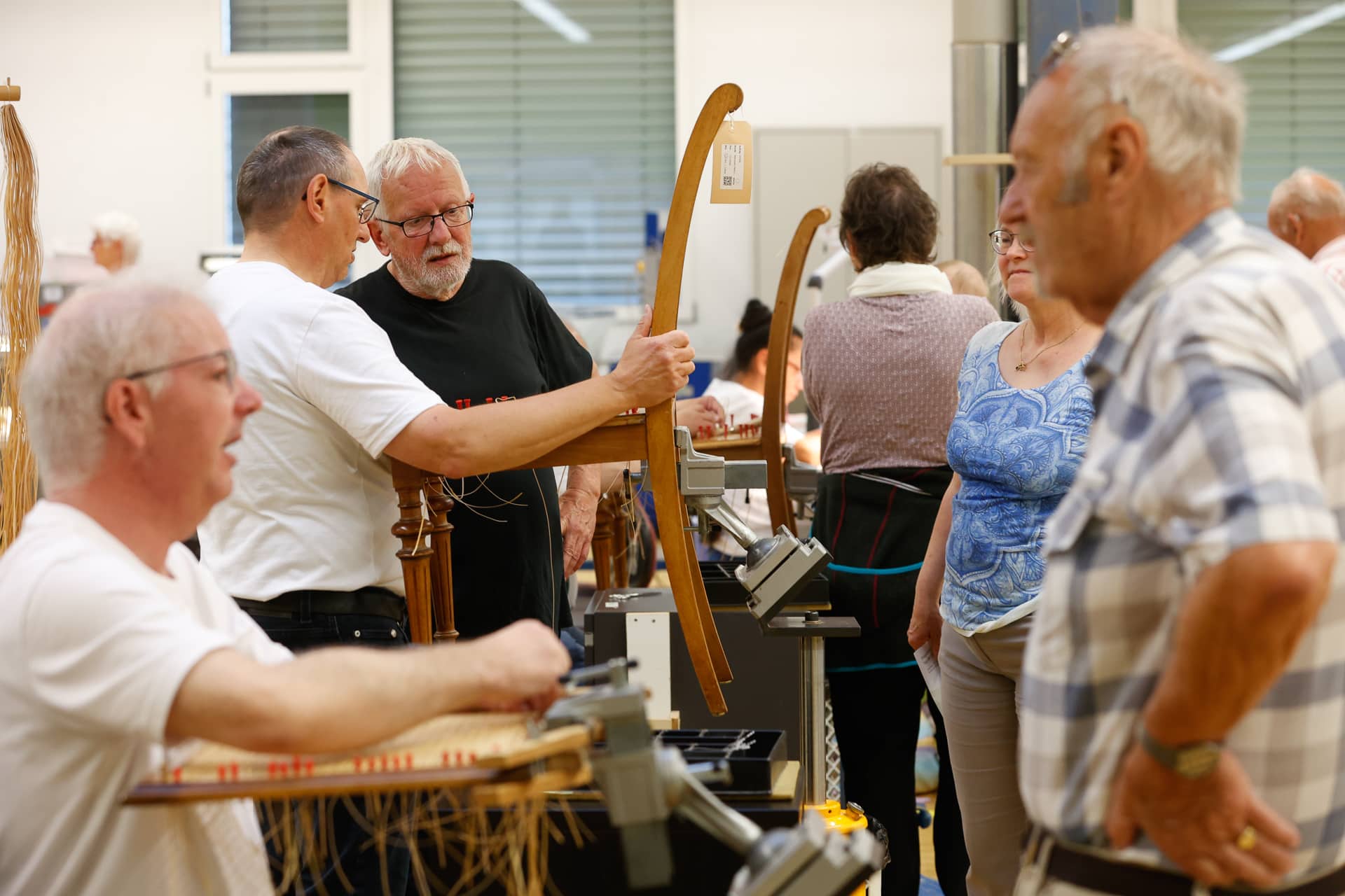 Eine Gruppe älterer Menschen hat sich zu einem Workshop versammelt, betrachtet Holzgegenstände und unterhält sich; ein Mann hält ein gebogenes Holzstück in der Hand, während die anderen zuschauen und darüber diskutieren.