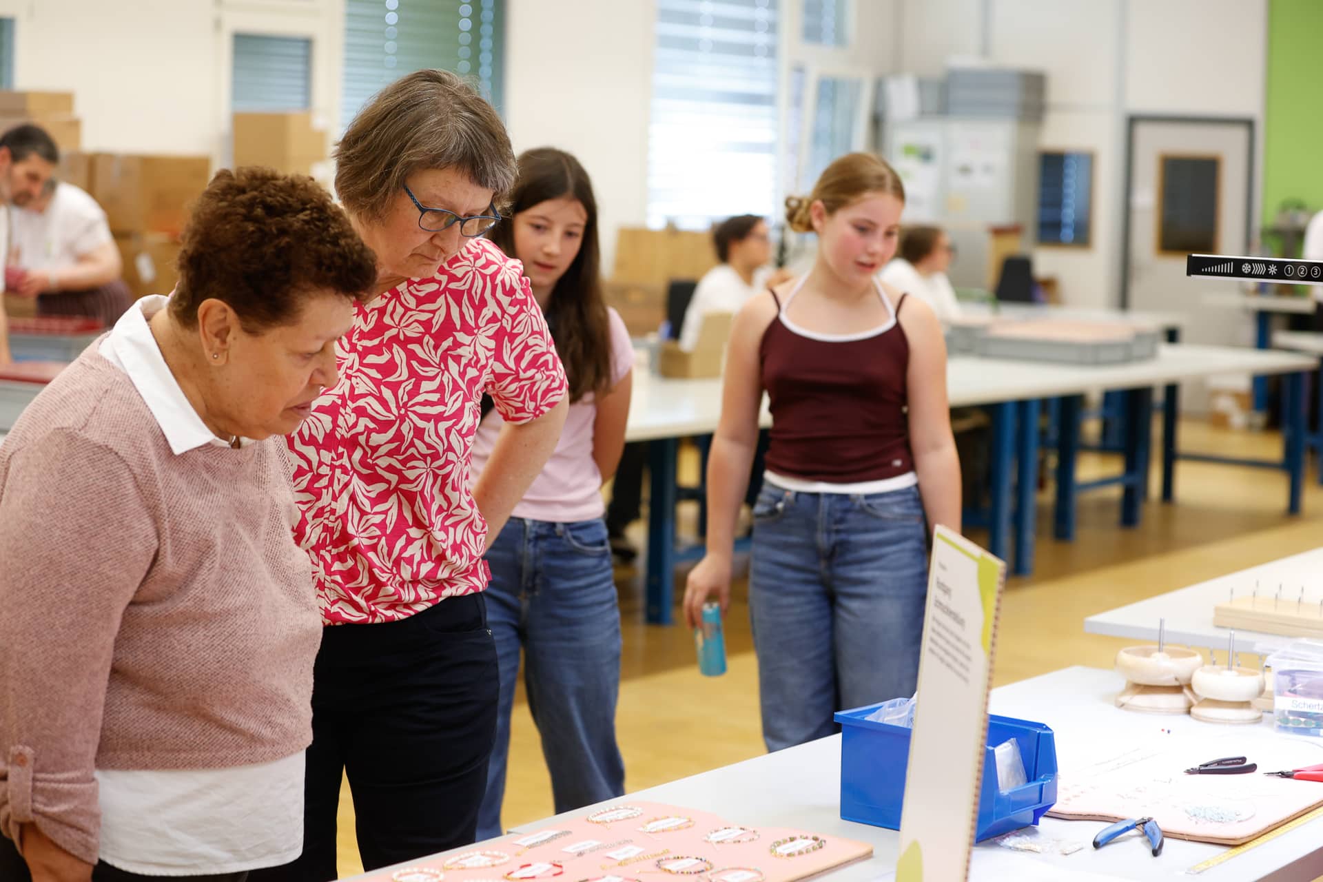 Zwei Frauen betrachten aufmerksam Gegenstände auf einem Tisch in einem hellen Raum, während zwei Mädchen daneben stehen. Im Hintergrund sind weitere Personen, Regale und Kisten zu sehen, was auf eine Werkstatt oder ein Klassenzimmer hindeutet.