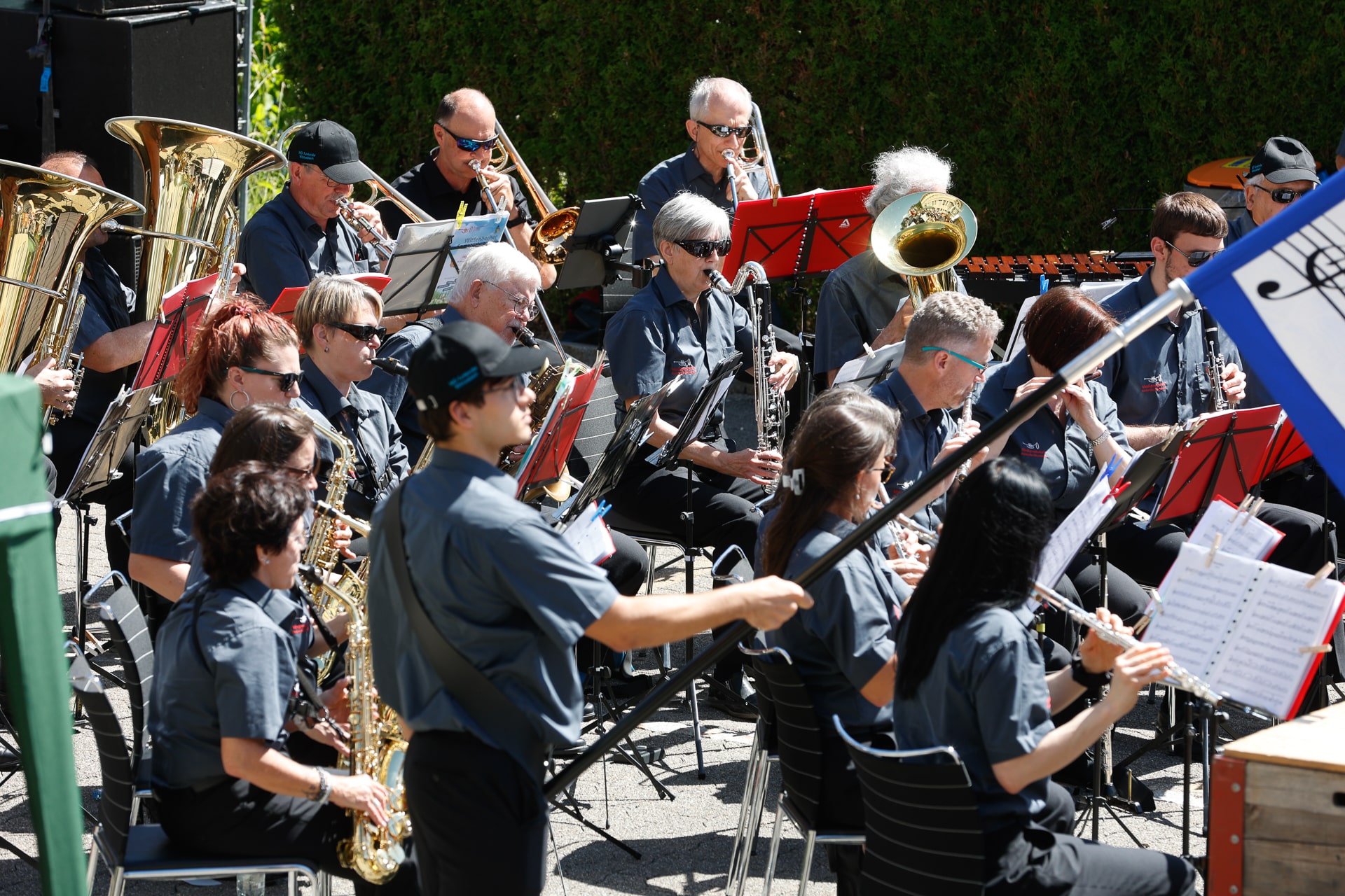 Eine große Musikgruppe, deren Mitglieder einheitliche graue Hemden tragen, spielt an einem sonnigen Tag im Freien verschiedene Blechblas-, Holzblas- und Schlaginstrumente, während sie in Reihen mit Notenständern sitzen.