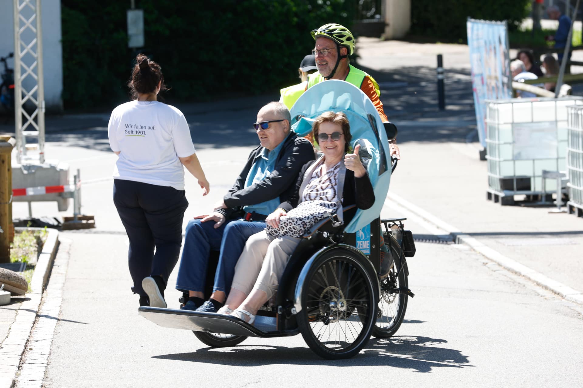Ein Mann tritt in die Pedale eines Rikscha-ähnlichen Fahrrads, auf dem zwei lächelnde ältere Fahrgäste sitzen, während eine Frau auf einer sonnigen Straße vor ihm hergeht. Die Fahrgäste sitzen bequem und scheinen die Fahrt zu genießen.