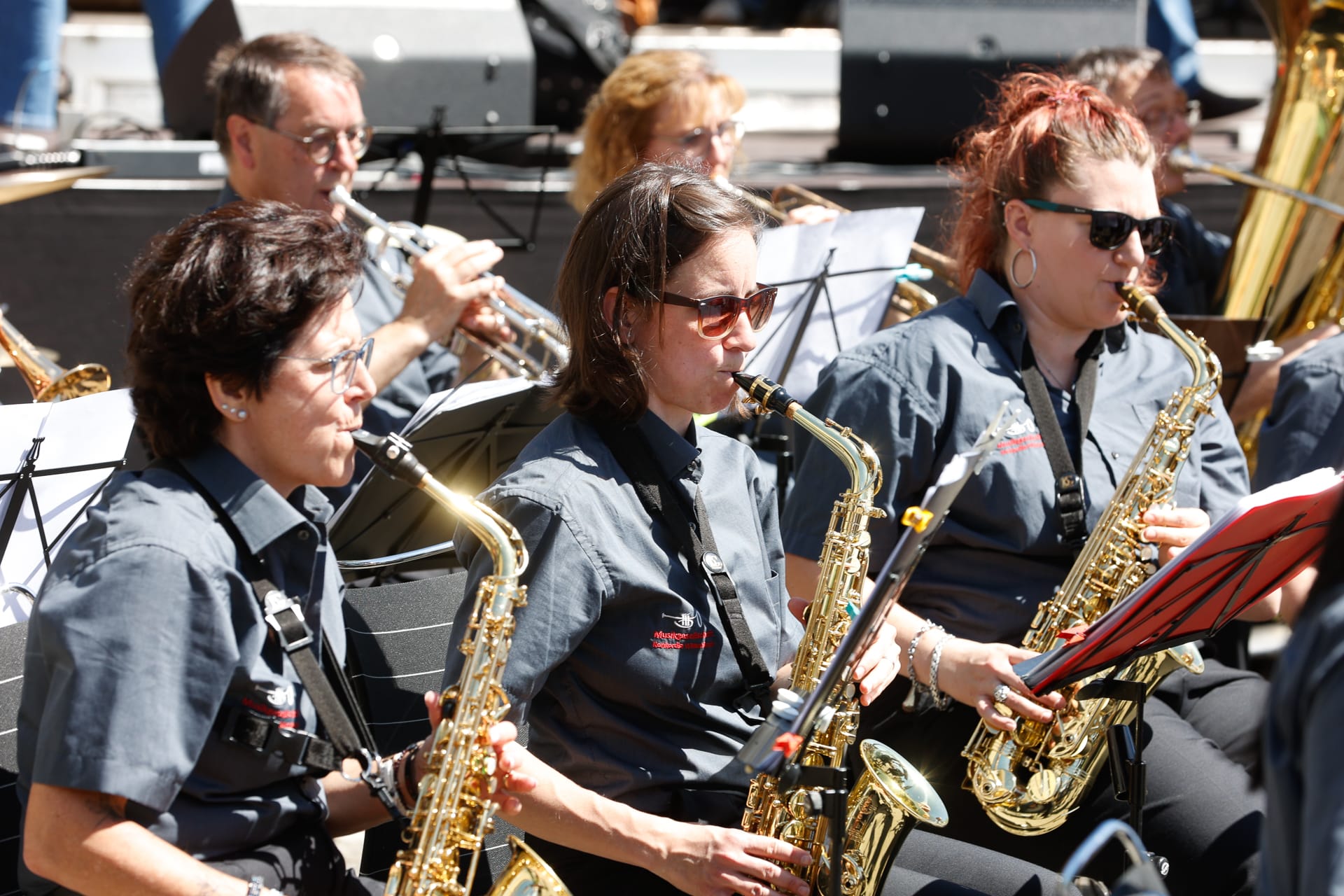 Eine Gruppe von Musikern, überwiegend Frauen, spielt im Freien bei strahlendem Sonnenschein Saxophon; sie tragen einheitliche graue Shirts und Sonnenbrillen und haben Noten vor sich liegen.