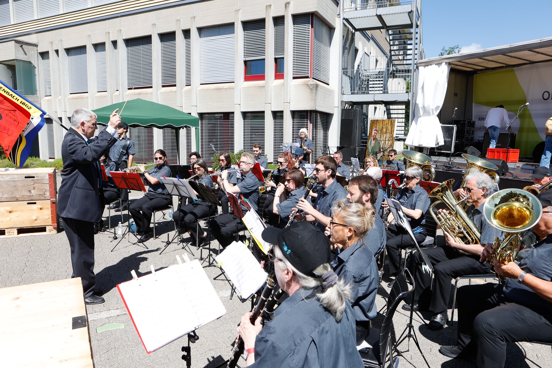 Ein Dirigent leitet ein großes Bläserorchester im Freien. Die Musiker sitzen in Reihen und spielen an einem sonnigen Tag vor einem modernen Gebäude. In der Nähe stehen Notenständer und ein grünes Vordach.