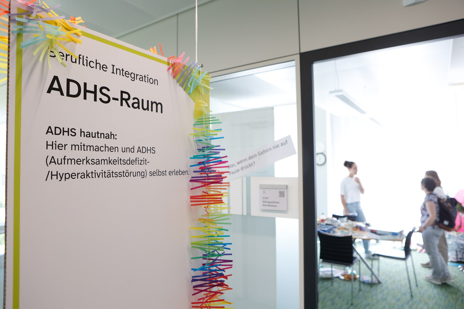 Am Eingang eines Raums hängt ein Schild mit der Aufschrift „ADHS-Raum“ und bunten Wäscheklammern. Durch die Glaswand sind mehrere Personen zu sehen, die sich in einem hellen, modernen Büroraum unterhalten.