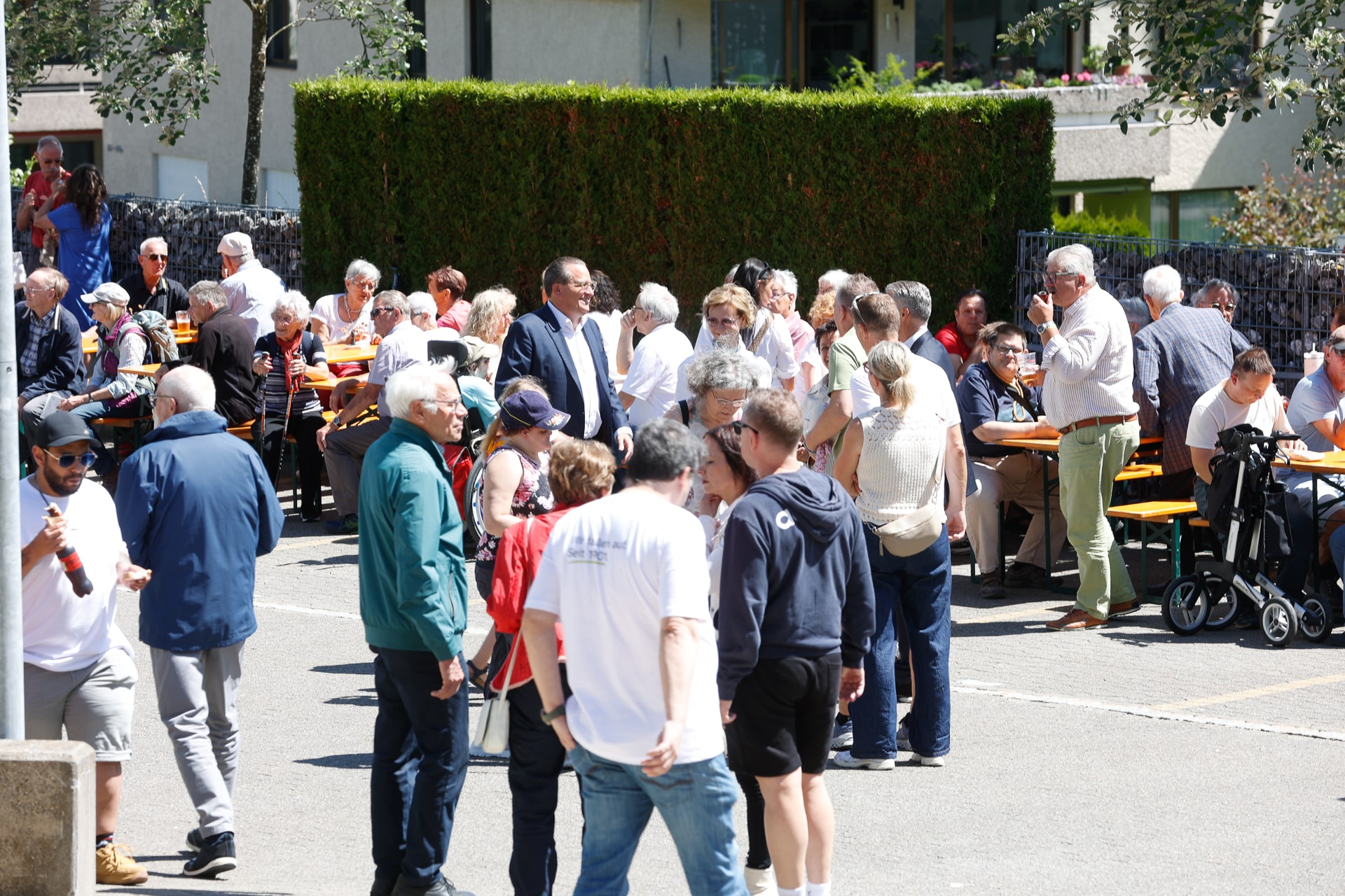 An einem sonnigen Tag versammelt sich eine große Gruppe von Menschen, hauptsächlich Erwachsene und Senioren, im Freien; sie sitzen und stehen an langen Tischen, unterhalten sich und genießen eine Veranstaltung in einem Innenhof.