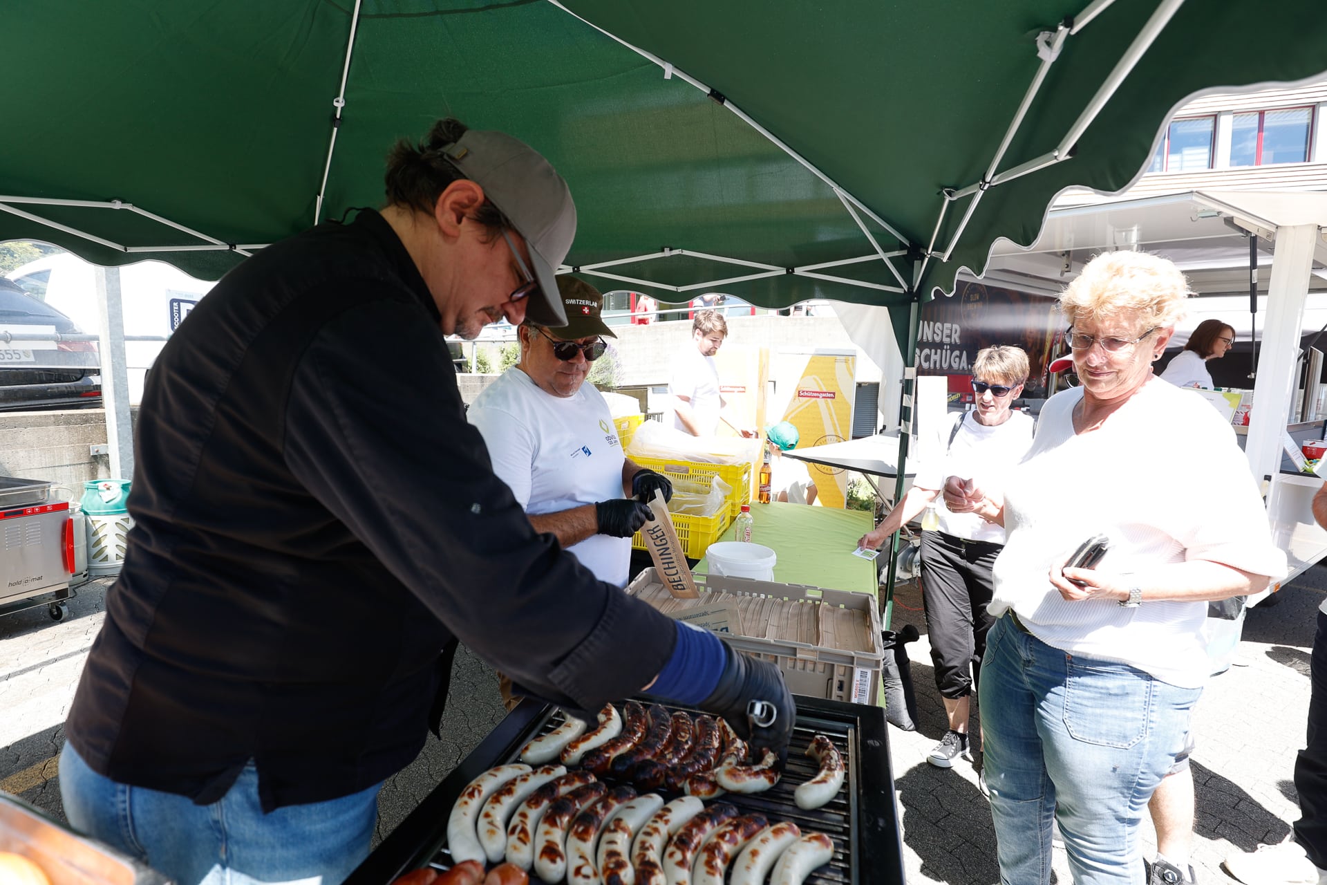 Auf einem Markt im Freien grillt jemand unter einem grünen Vordach Würstchen, während ein paar Leute in der Nähe stehen, lächeln und darauf warten, bedient zu werden. Im Hintergrund sind verschiedene Vorräte und Behälter zu sehen.