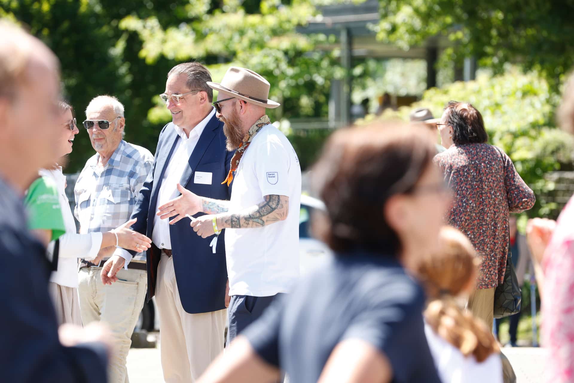Eine Gruppe von Menschen unterhält sich an einem sonnigen Tag im Freien, im Hintergrund sind Bäume zu sehen. Einige tragen Freizeitkleidung und Sonnenbrillen, während andere formeller gekleidet sind. Die Szene wirkt lebhaft und gesellig.