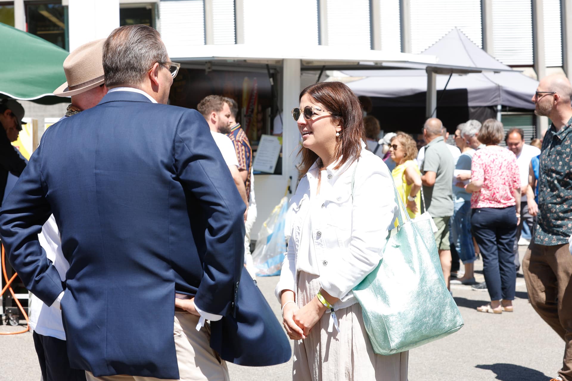 Bei einer Veranstaltung im Freien an einem sonnigen Tag unterhalten sich mehrere Menschen und lächeln. Im Hintergrund mischen sich weitere Personen in der Nähe von Zelten und Ständen unter die Menge, was an die Atmosphäre eines Marktes oder Festes erinnert.