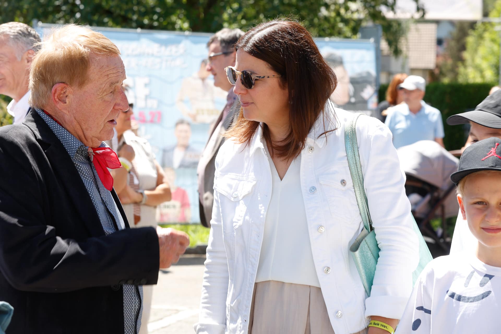 Ein älterer Mann in Anzug und rotem Schal unterhält sich bei einer Veranstaltung im Freien mit einer Frau, die eine Sonnenbrille und eine weiße Jacke trägt; im Hintergrund sind Menschen und ein verschwommenes Transparent zu sehen. Ein Junge mit einer Mütze steht lächelnd daneben.