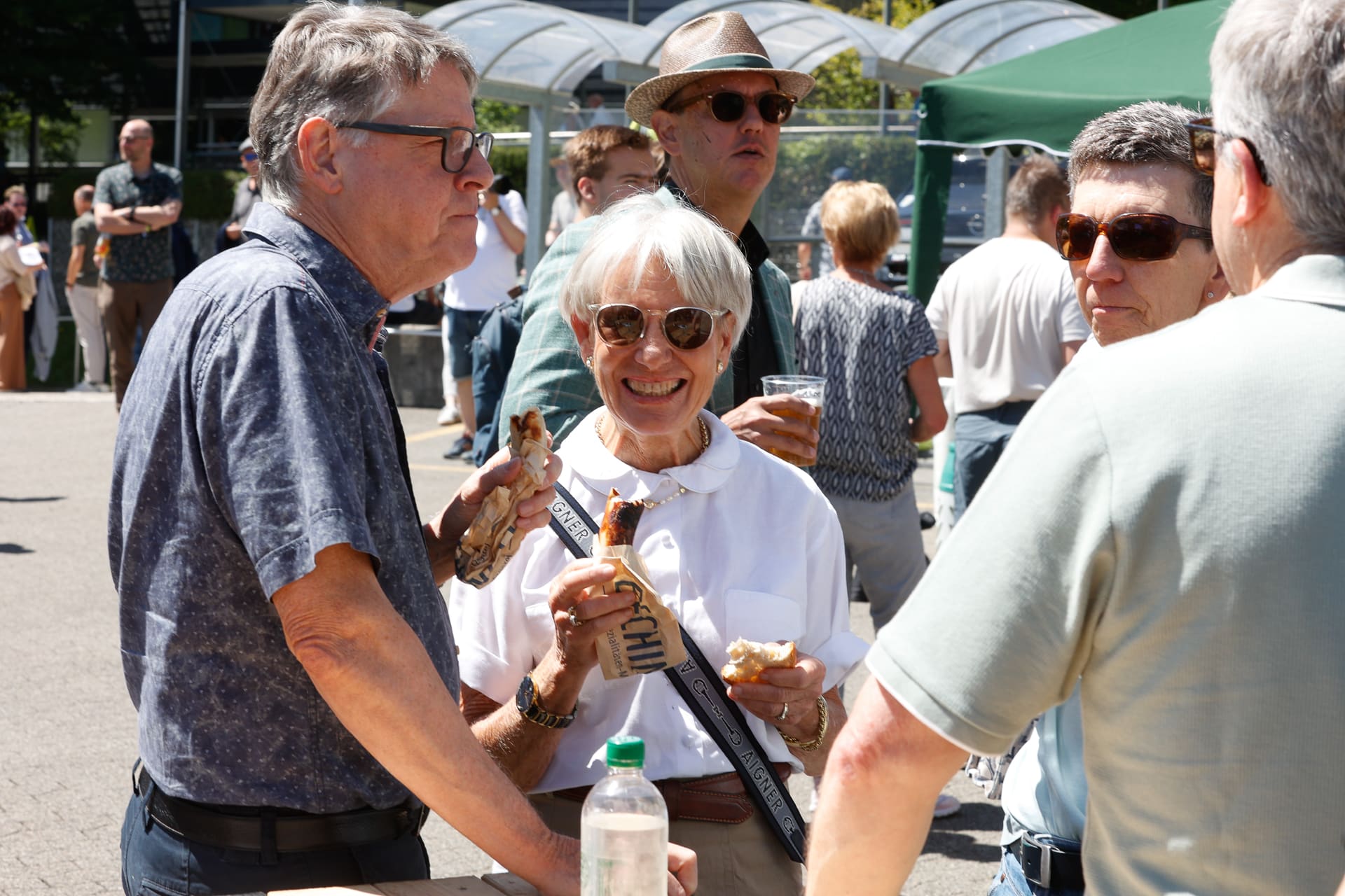 Eine Gruppe älterer Menschen steht bei einer Veranstaltung im Freien in der Sonne, unterhält sich und isst etwas. Eine Frau in der Mitte lächelt, während sie einen Hotdog in der Hand hält. Die Menschen um sie herum tragen Sonnenbrillen und Freizeitkleidung.