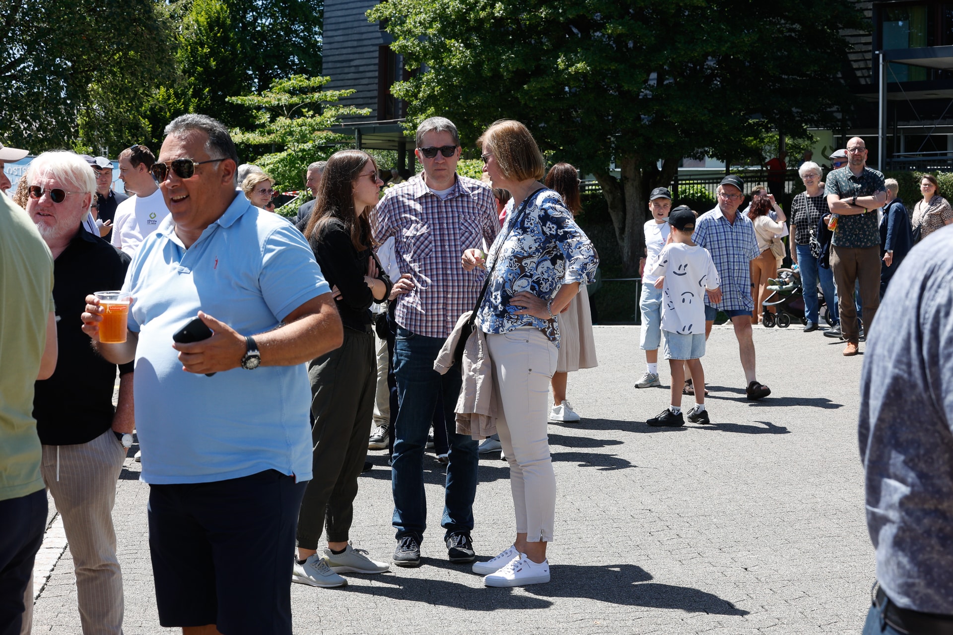 Eine Gruppe von Menschen steht draußen an einem sonnigen Ort; einige unterhalten sich und halten Getränke in der Hand, während andere in einer langen Schlange warten. Die meisten sind leger gekleidet, und im Hintergrund spenden Bäume Schatten.