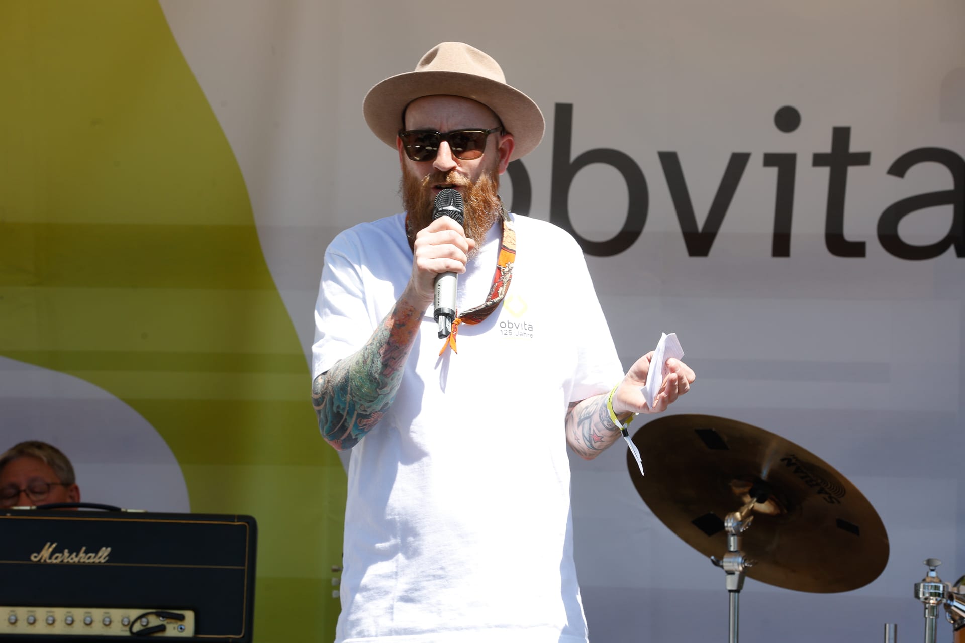 Eine bärtige Person mit Hut und Sonnenbrille spricht auf einer Außenbühne in ein Mikrofon und hält dabei ein Blatt Papier in der Hand. Im Hintergrund sind Musikinstrumente zu sehen, darunter ein Schlagzeug und ein Verstärker.
