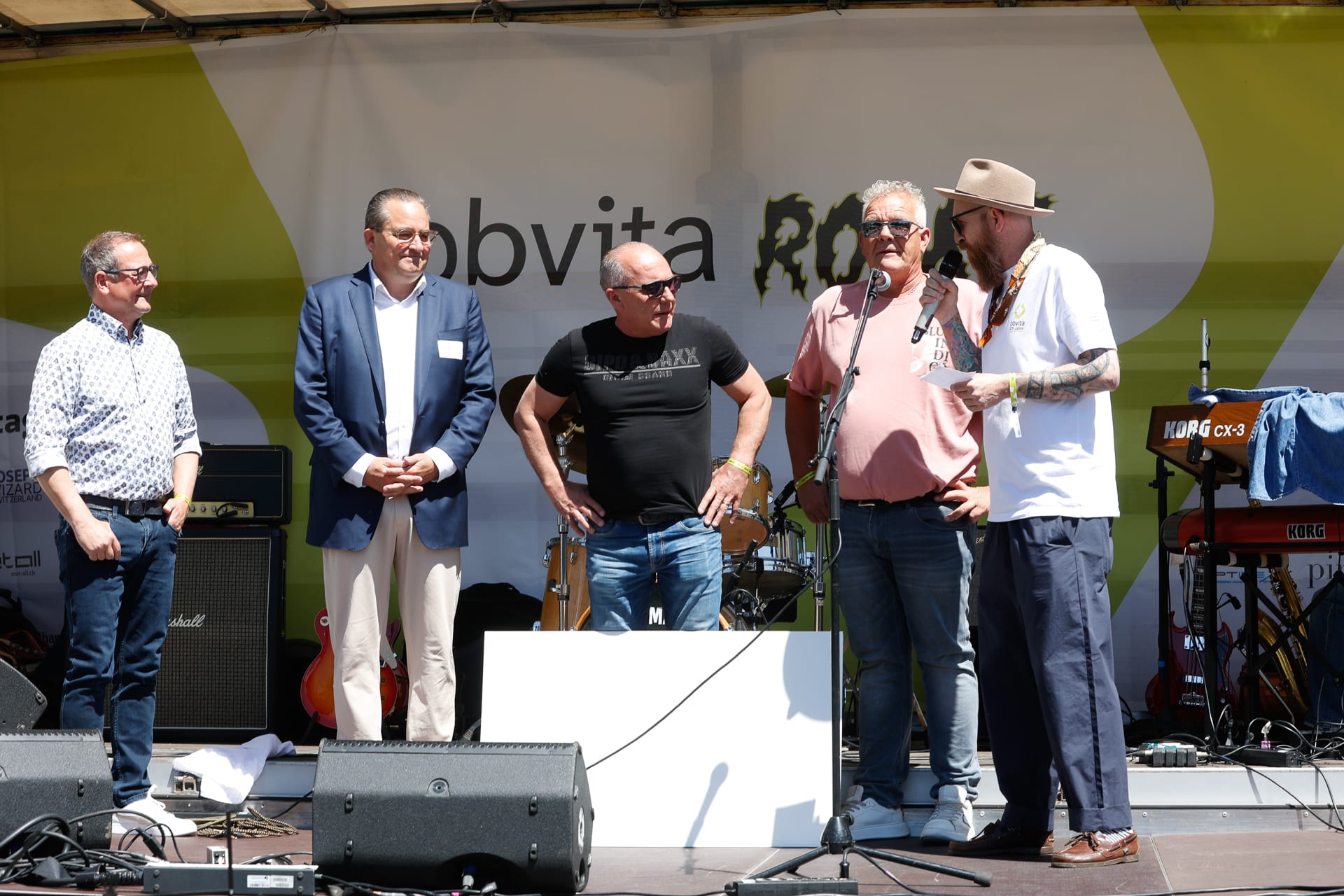 Fünf Männer stehen auf einer Freiluftbühne, im Hintergrund sind Musikinstrumente zu sehen; ein Mann spricht in ein Mikrofon, während die anderen zuhören. Auf der Kulisse sind die Wörter „obvita“ und „ROCK“ zu sehen.