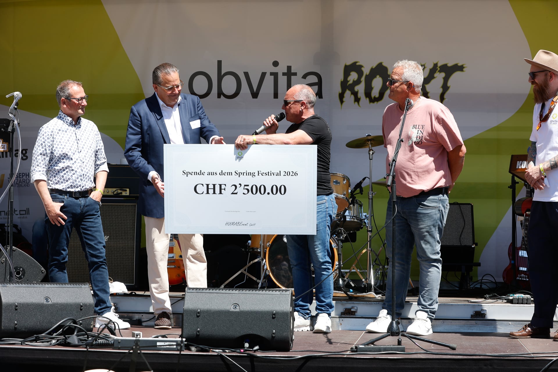 Fünf Männer stehen auf einer Freiluftbühne. Ein Mann überreicht einem anderen einen großen Scheck über 2.500,00 CHF. Im Hintergrund sind Musikinstrumente und ein gelb-weißes Transparent zu sehen.