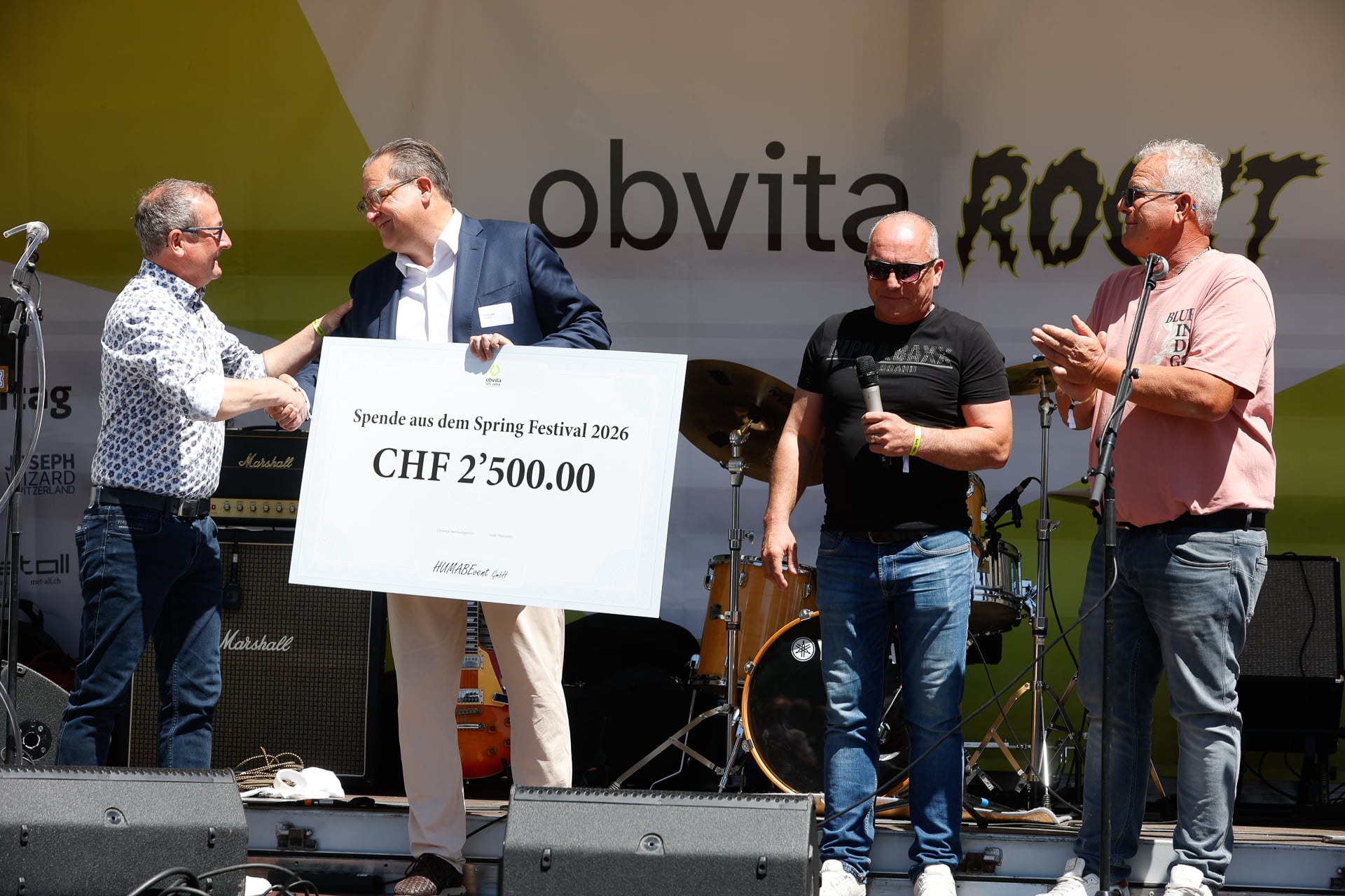 Vier Männer stehen auf der Bühne; einer überreicht einem anderen einen großen Scheck über 2'500 CHF. Zwei weitere Männer stehen neben ihnen, einer klatscht. Im Hintergrund sind Musikinstrumente und ein Festbanner zu sehen.