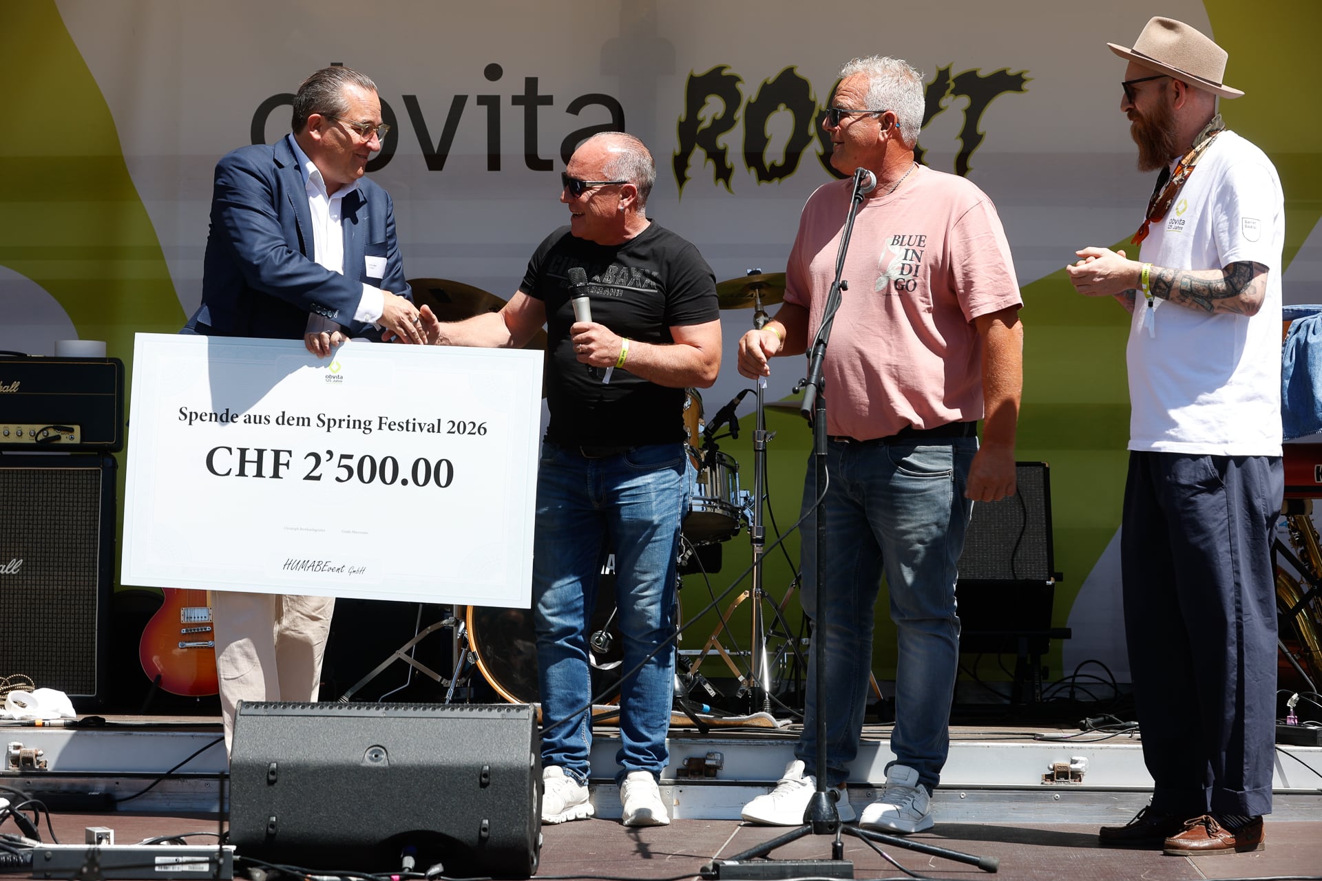 Vier Männer stehen auf der Bühne. Einer überreicht einem anderen einen großen Scheck über 2.500,00 CHF. Sie lächeln und halten Mikrofone in der Hand. Im Hintergrund sind Musikinstrumente und ein Transparent zu sehen.