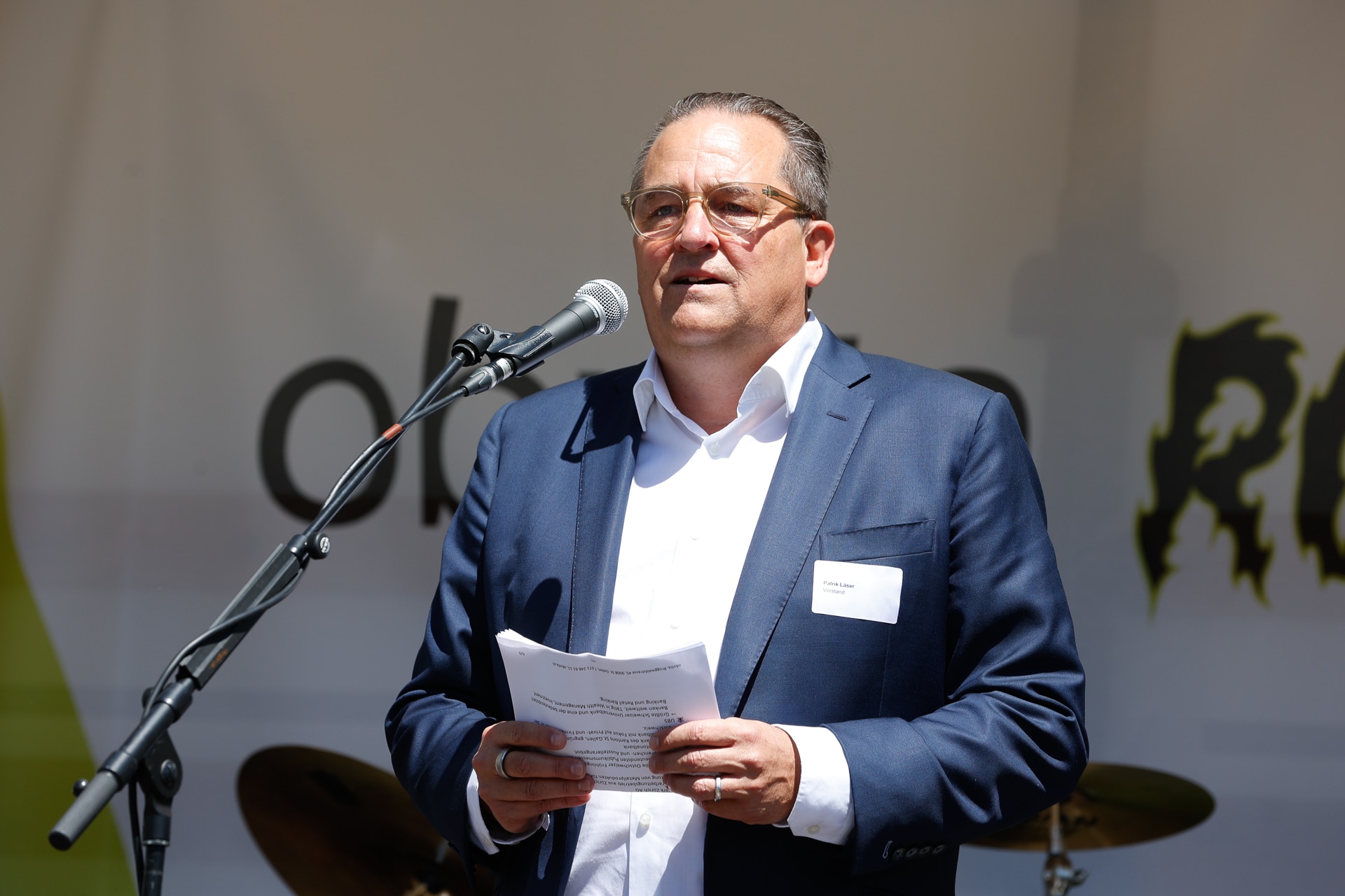 Ein Mann in einem marineblauen Anzug und mit Brille steht vor einem Mikrofon, hält Papiere in der Hand und spricht im Freien. Im Hintergrund ist ein Schlagzeug zu sehen, und hinter ihm sind Teile der Veranstaltungsbeschilderung zu erkennen.