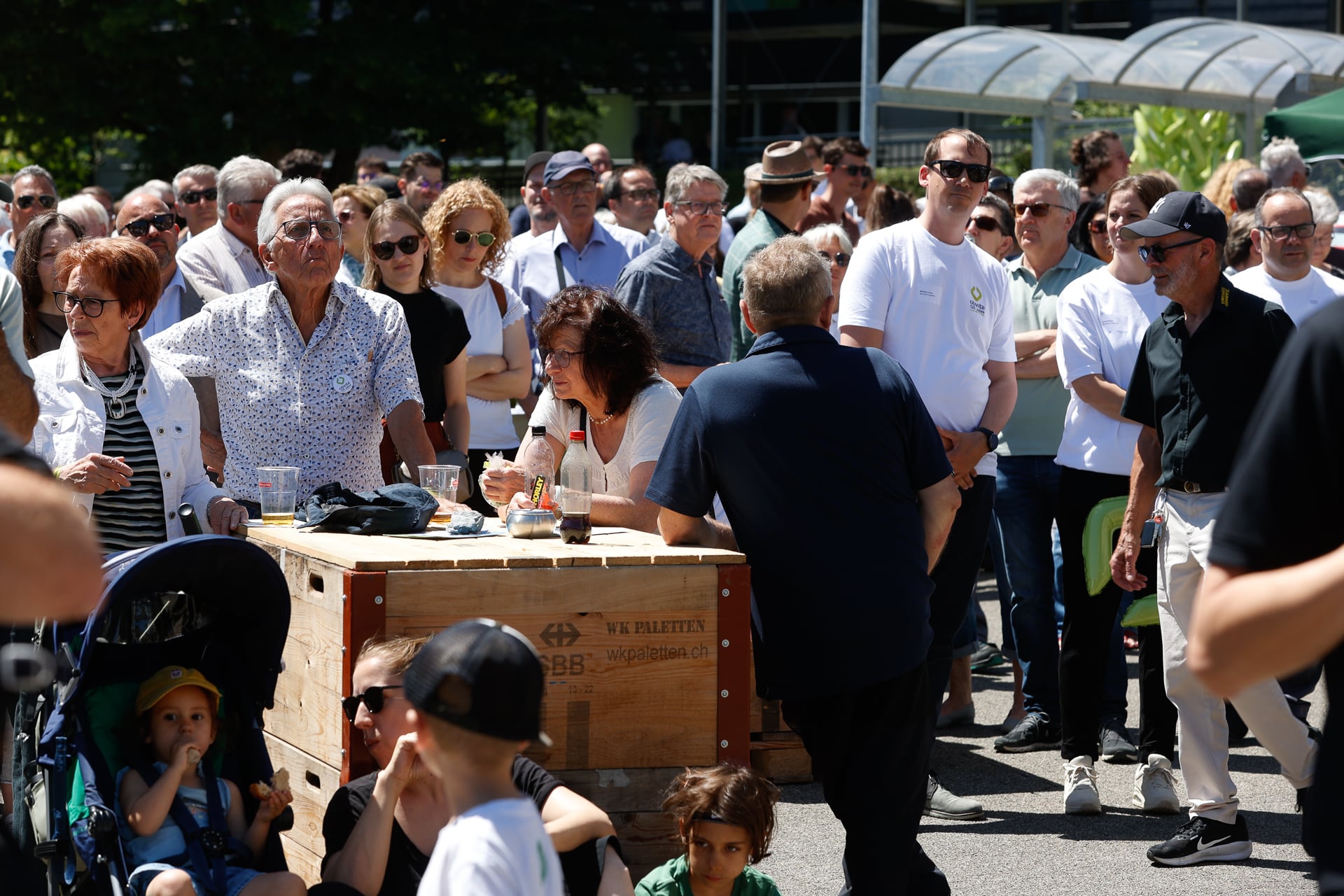 An einem sonnigen Tag versammelt sich eine große Gruppe von Menschen, darunter Erwachsene und Kinder, im Freien. Einige stehen, während andere sitzen oder an einem Holztisch essen. Die Atmosphäre wirkt gesellig und lebhaft.